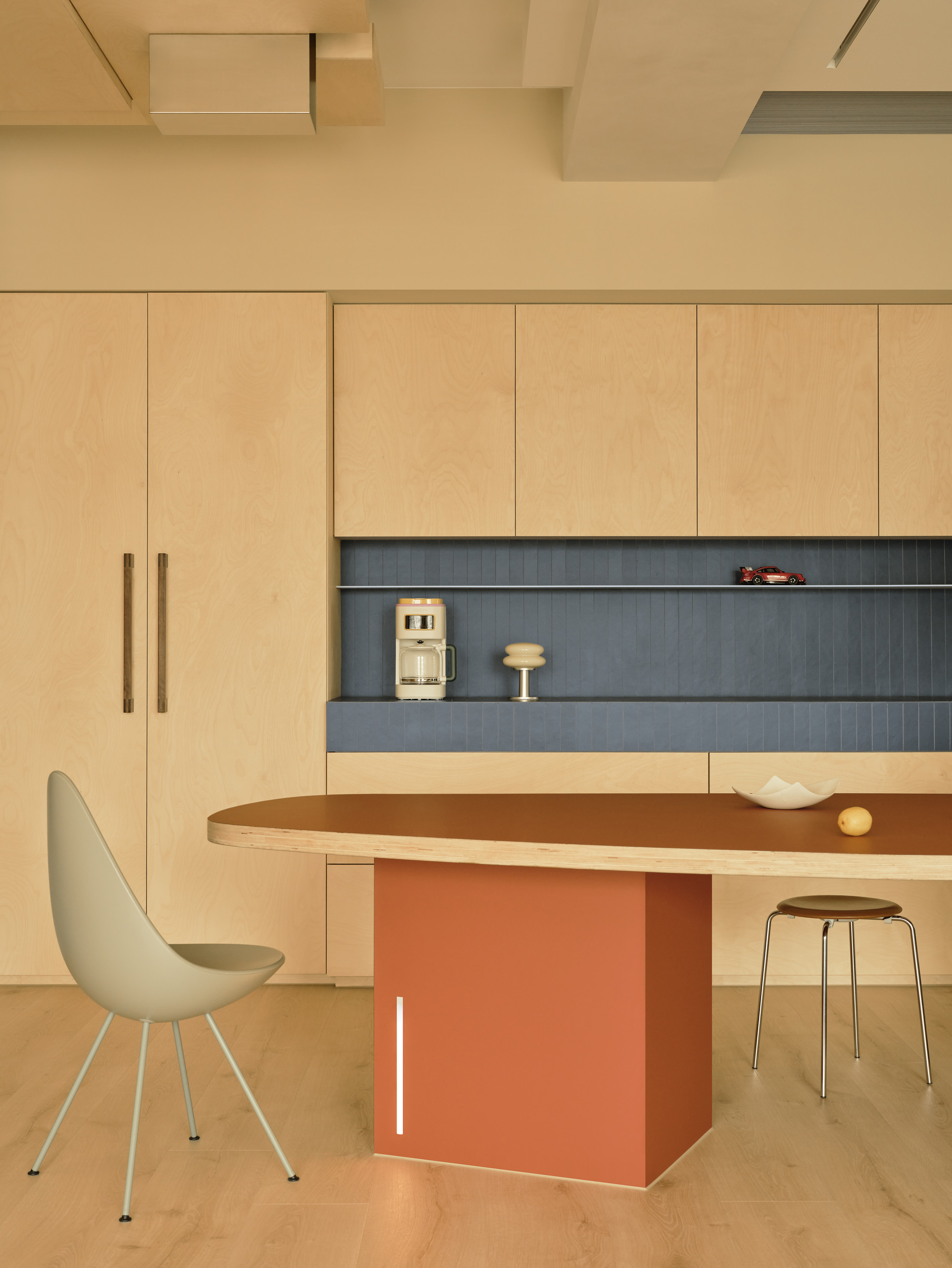 Rust-colored table base and teardrop chair in front of plywood cabinets and a blue tiled shelf with a coffee maker