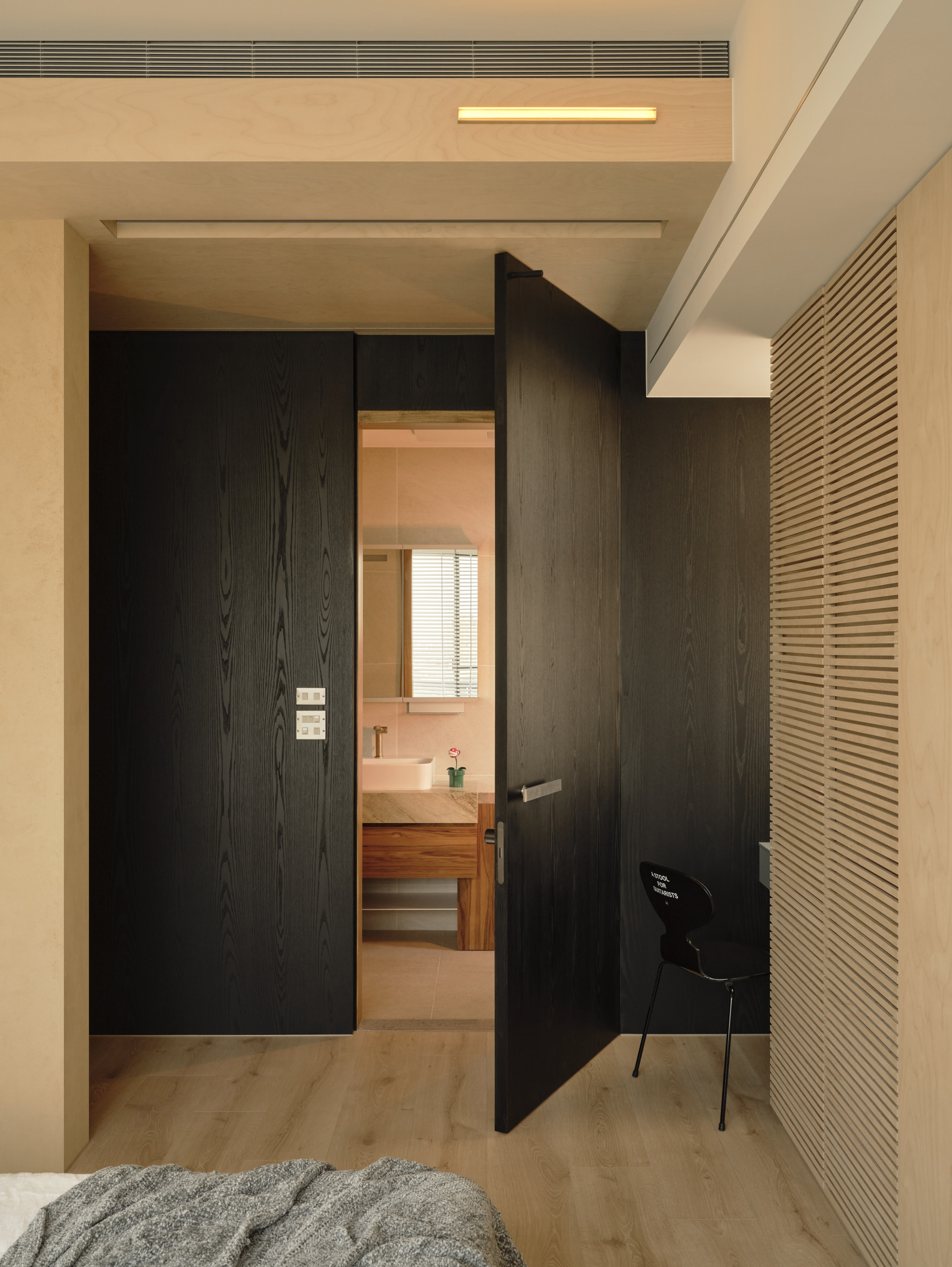 Black-stained timber pivot door open onto a bathroom, with a slatted plywood wall and a black chair beside it