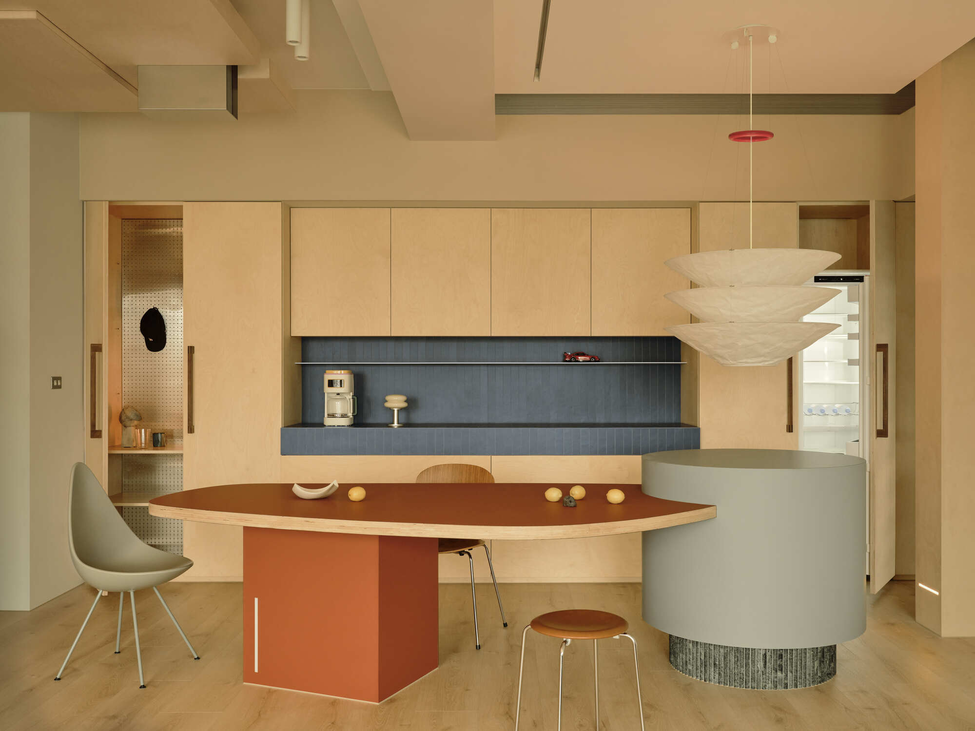 Kitchen wall with plywood doors opened to reveal a pegboard pantry and a refrigerator beside the round gray island