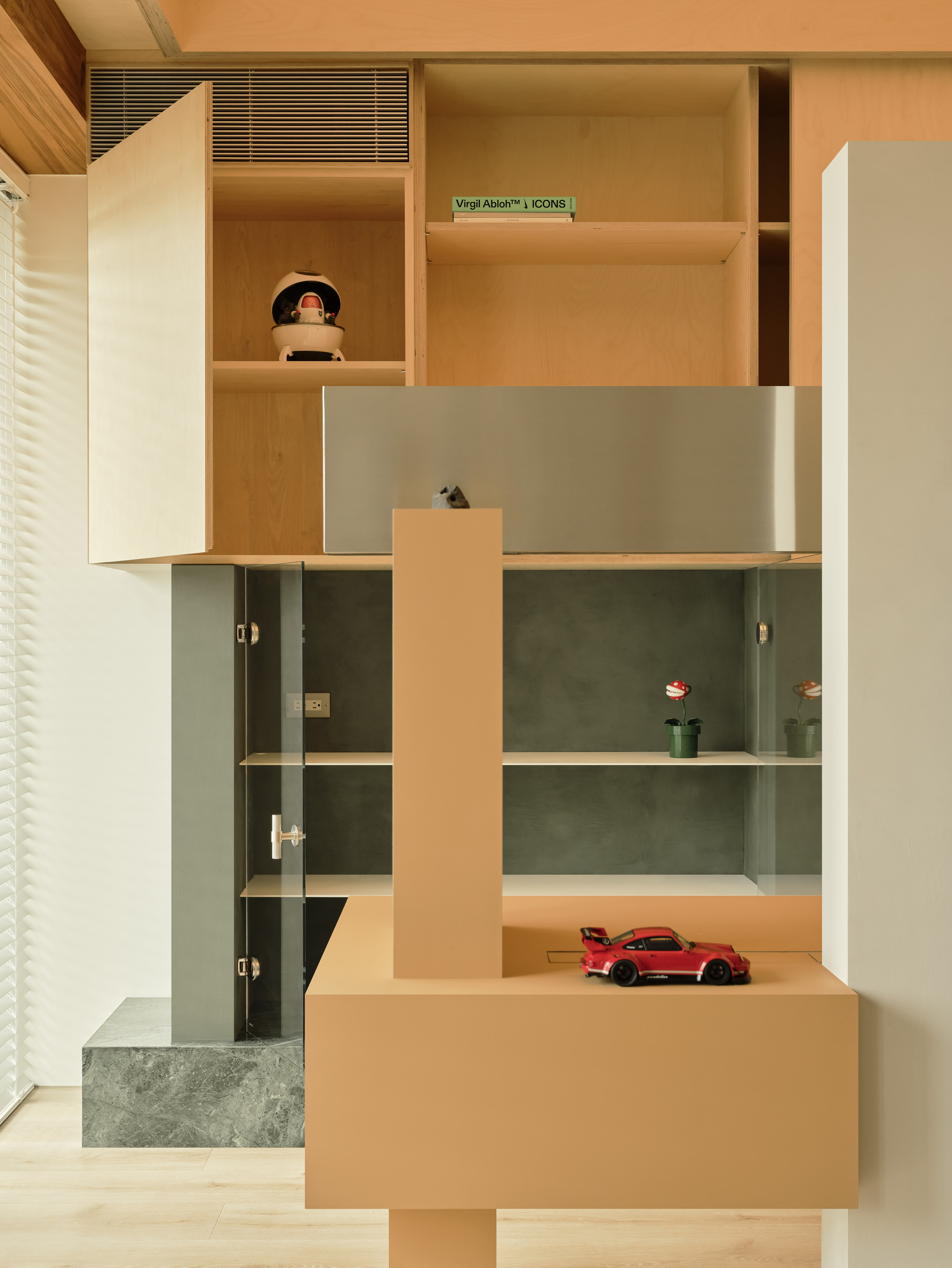 Stacked plywood shelving with an open cabinet door above a peach-colored block holding a red toy car, beside a green stone plinth