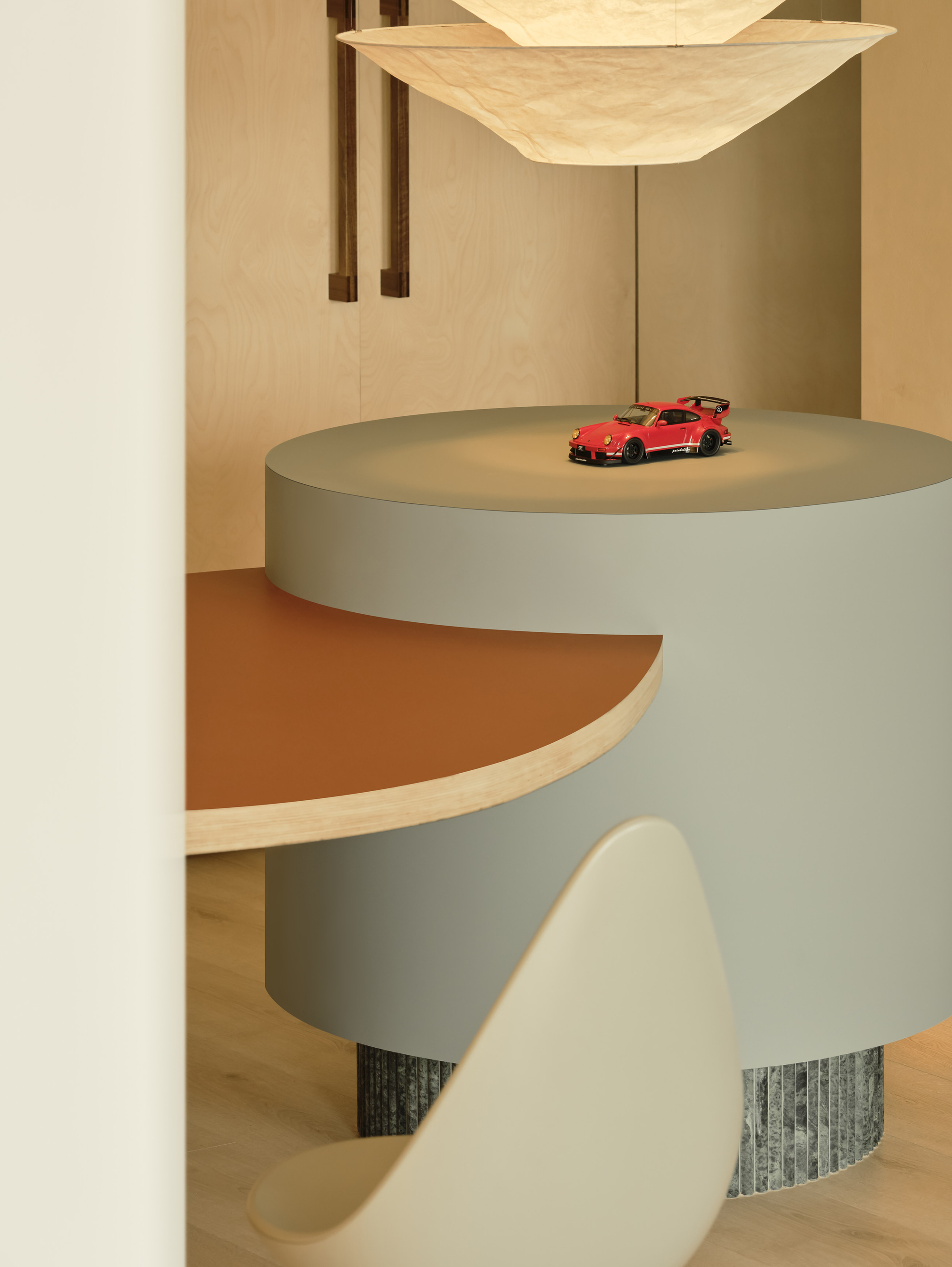 Round grey pedestal table with a rust-colored half-disc top, a red toy car on top and a paper pendant lamp above