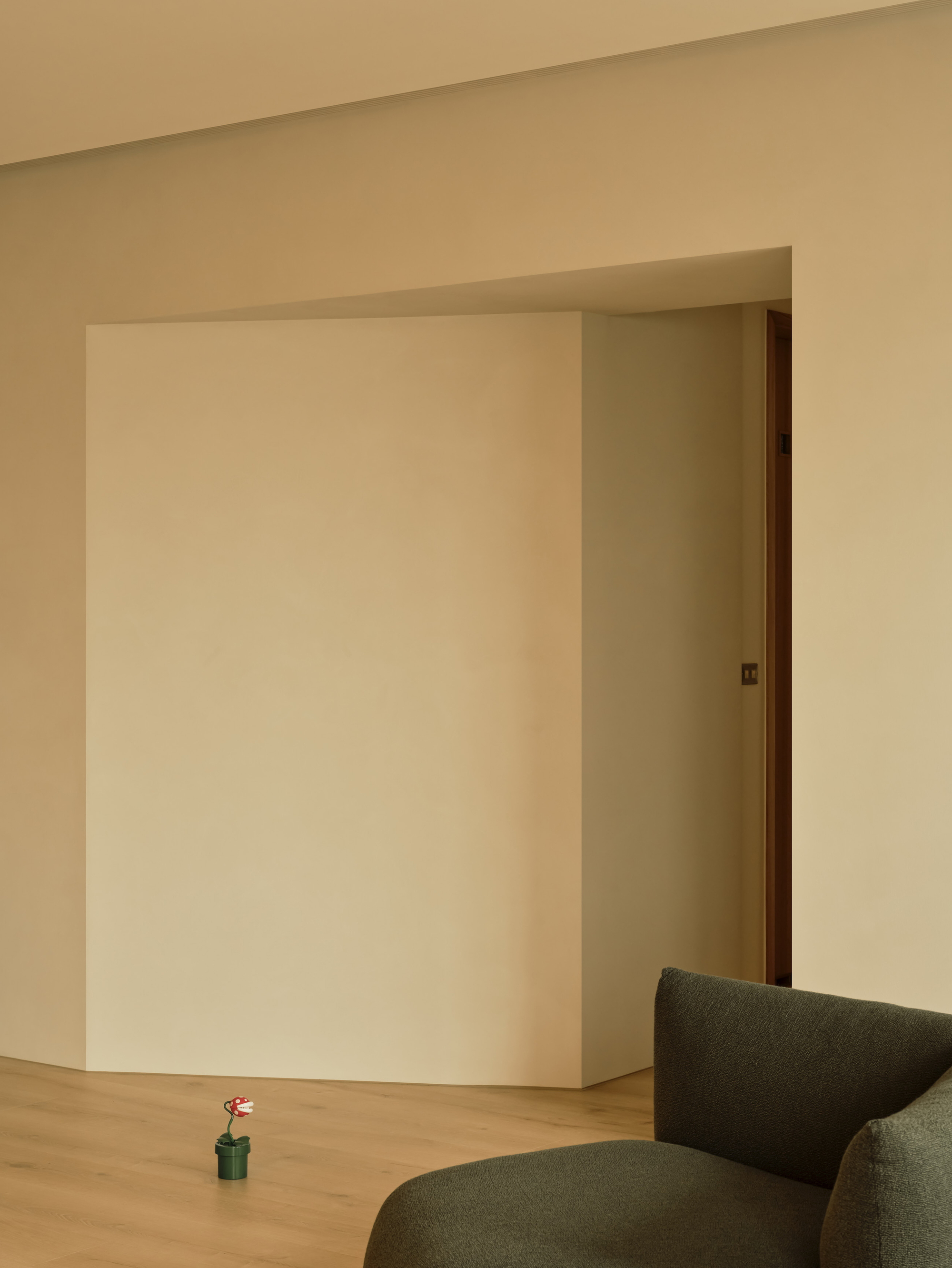 Angled cream plaster wall panel beside a doorway, with a small potted toy plant on the wood floor and a green sofa edge