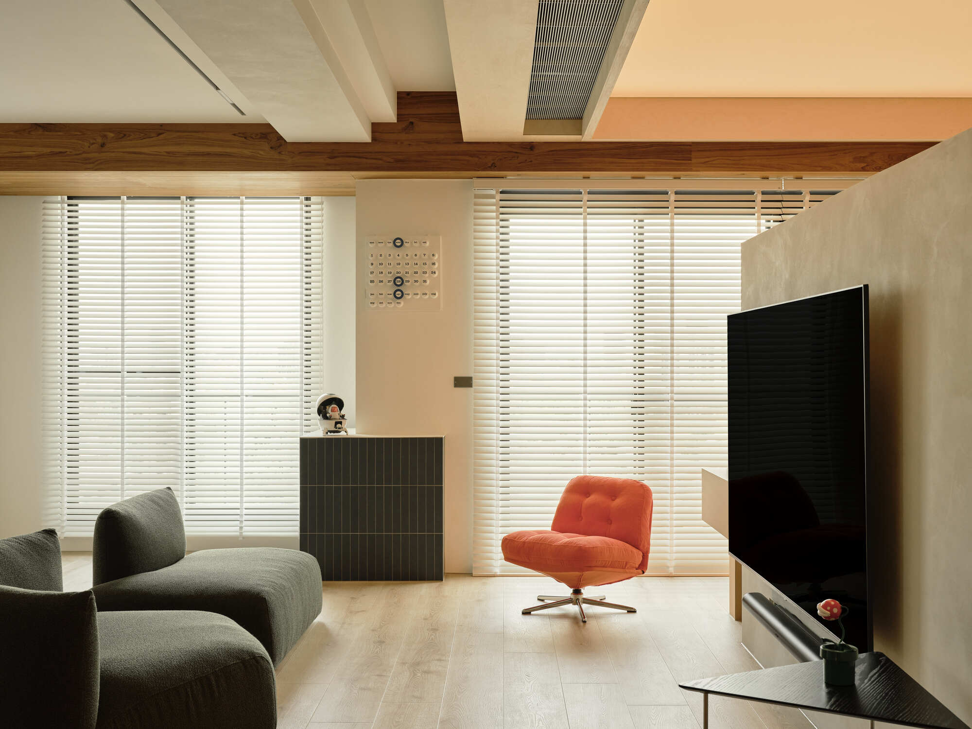 Living area with an orange swivel chair before white slatted blinds, a timber ceiling beam and olive sofas in soft daylight