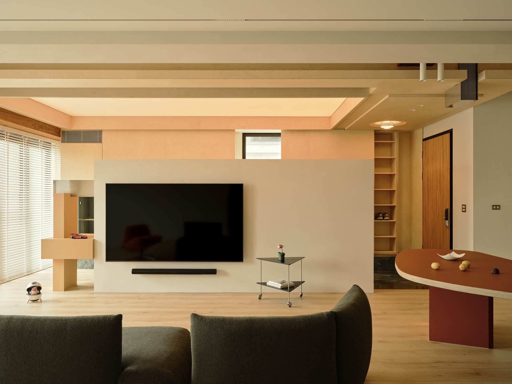 Freestanding plaster wall with a wall-mounted television, plywood ceiling beams, open shoe shelves and a rust-colored table edge in warm light