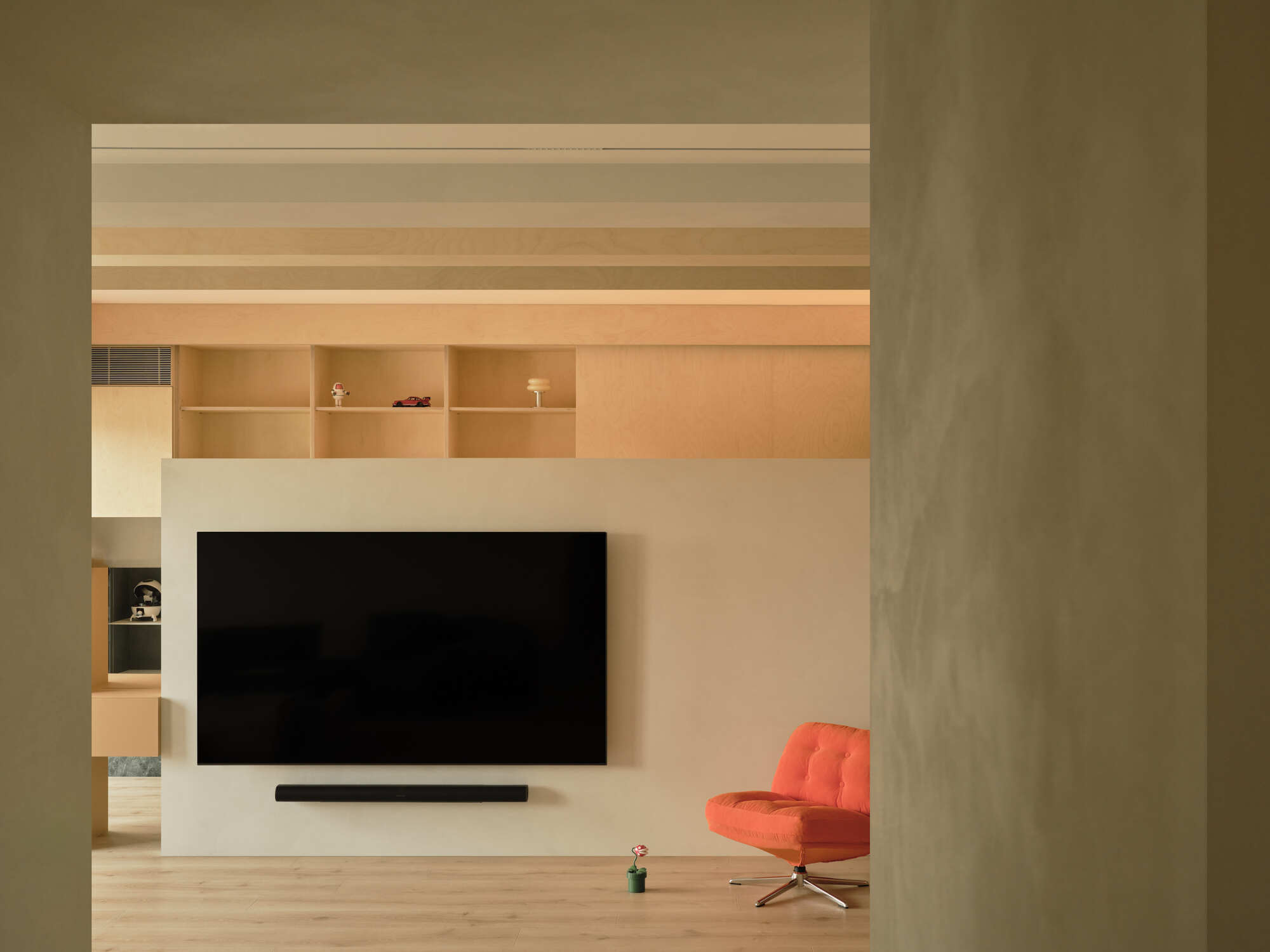 View through a doorway to the television wall topped with plywood cubbies, an orange chair standing at its foot