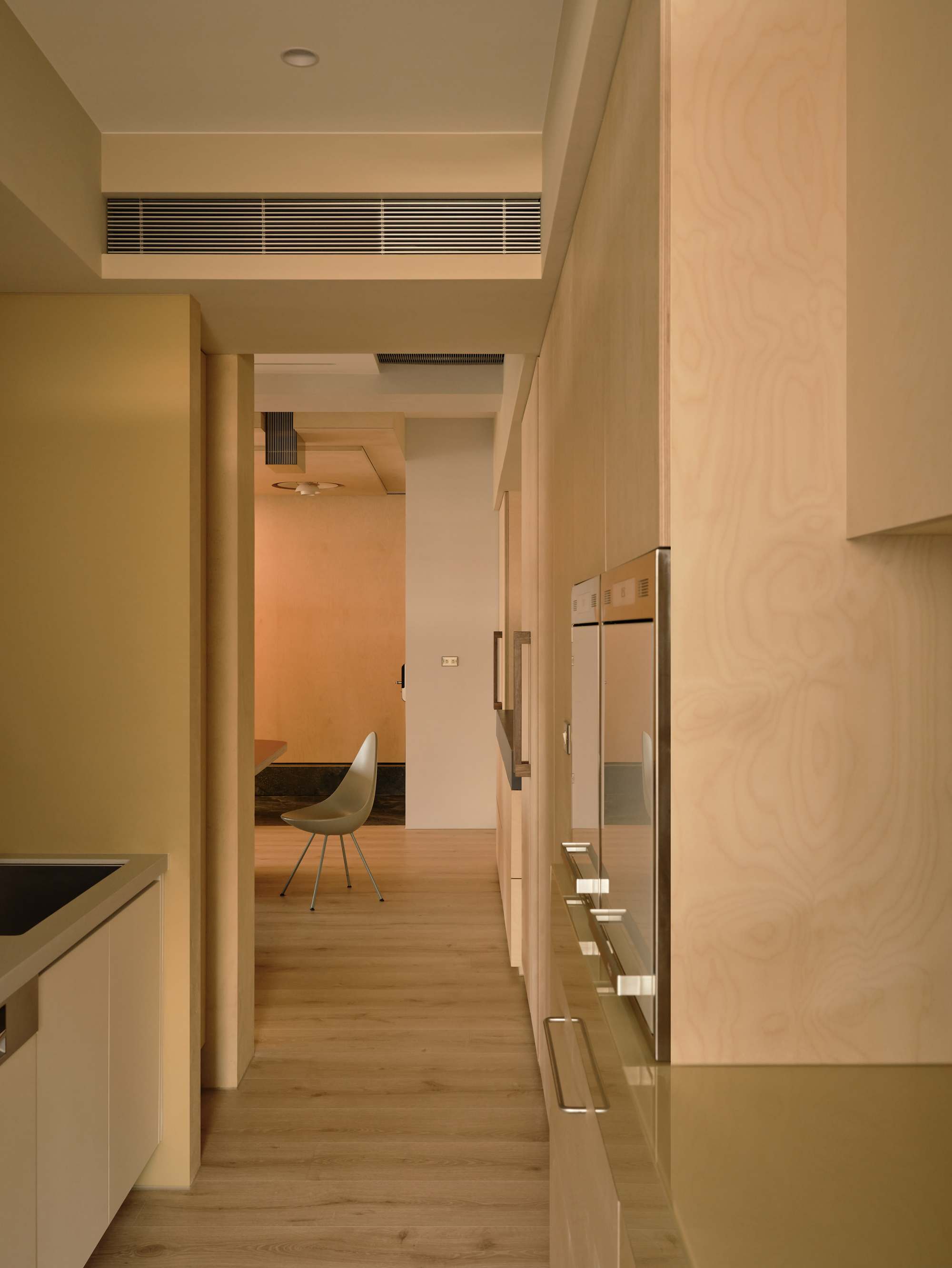 Narrow corridor lined with birch plywood cabinetry and built-in ovens, leading to a molded chair in a warm peach-lit room