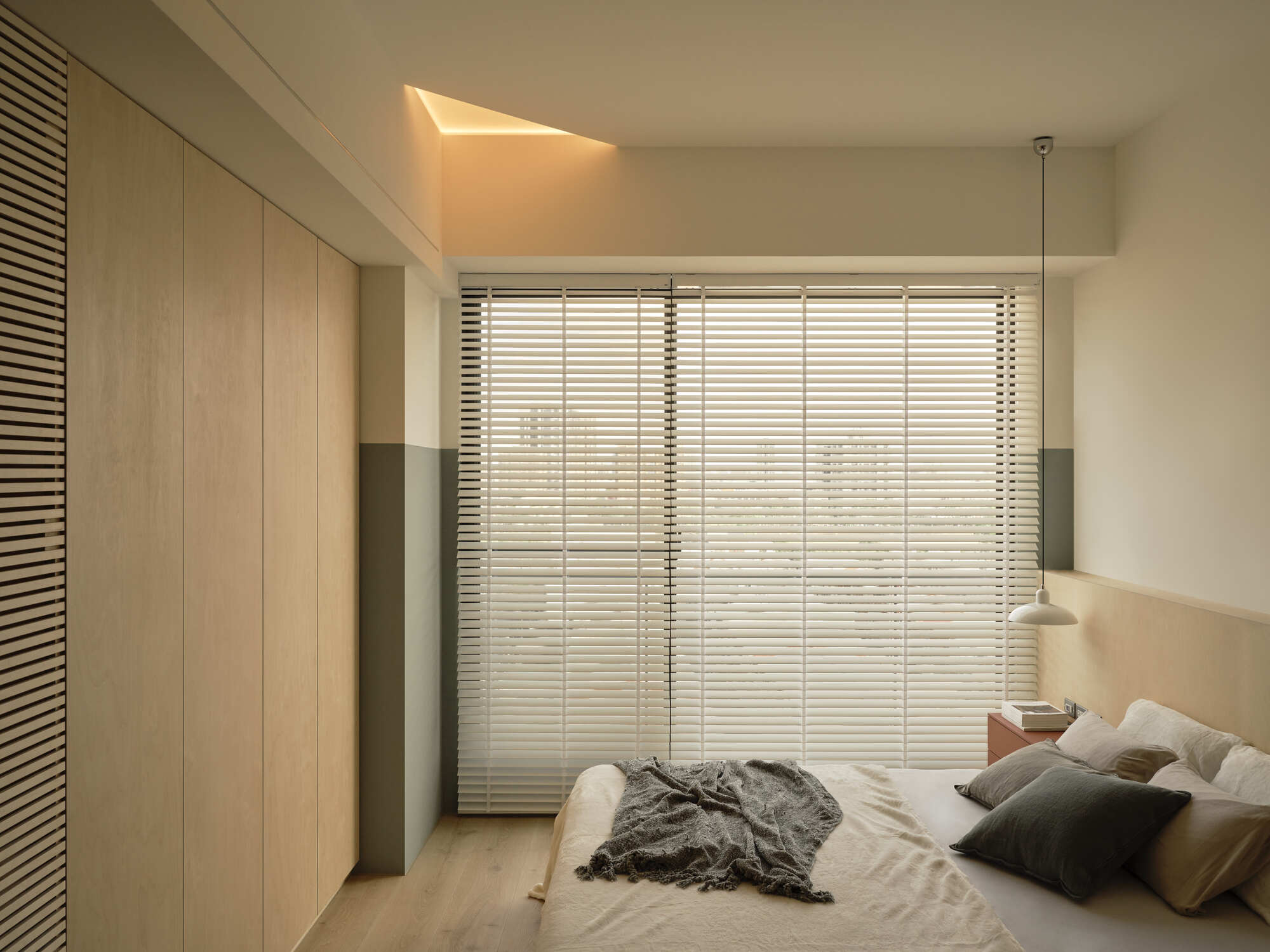 Bedroom with a wall of pale wood wardrobe doors facing a blinded window, a concealed skylight glowing at the ceiling edge