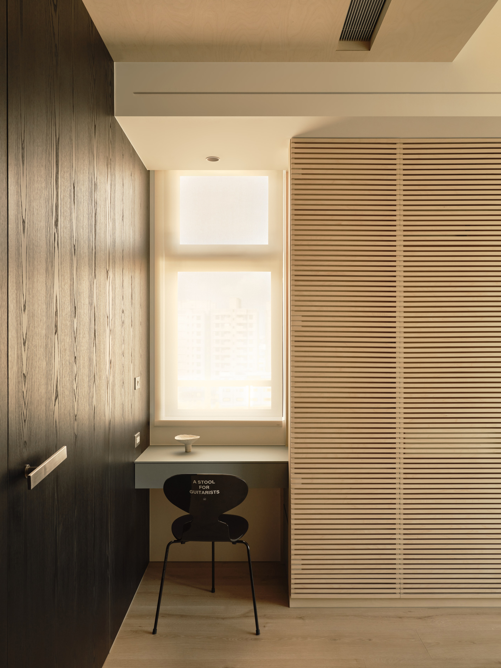 Built-in desk and black chair beside a window, between a dark wood-veneer wall and a slatted wardrobe in soft daylight