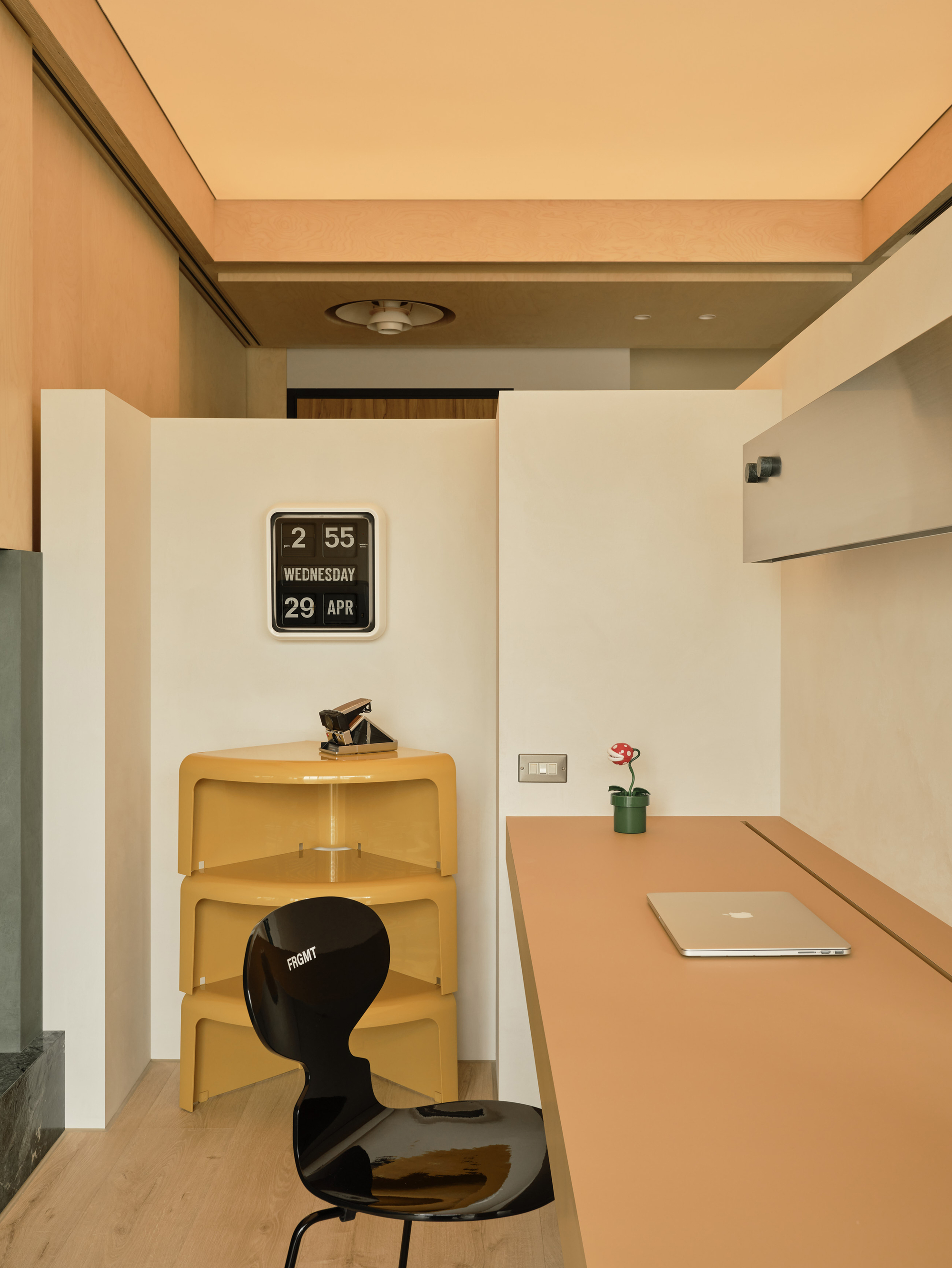 Orange desk with laptop and small plant beside stacked yellow shelves, a flip clock and a glossy black chair under a lit ceiling