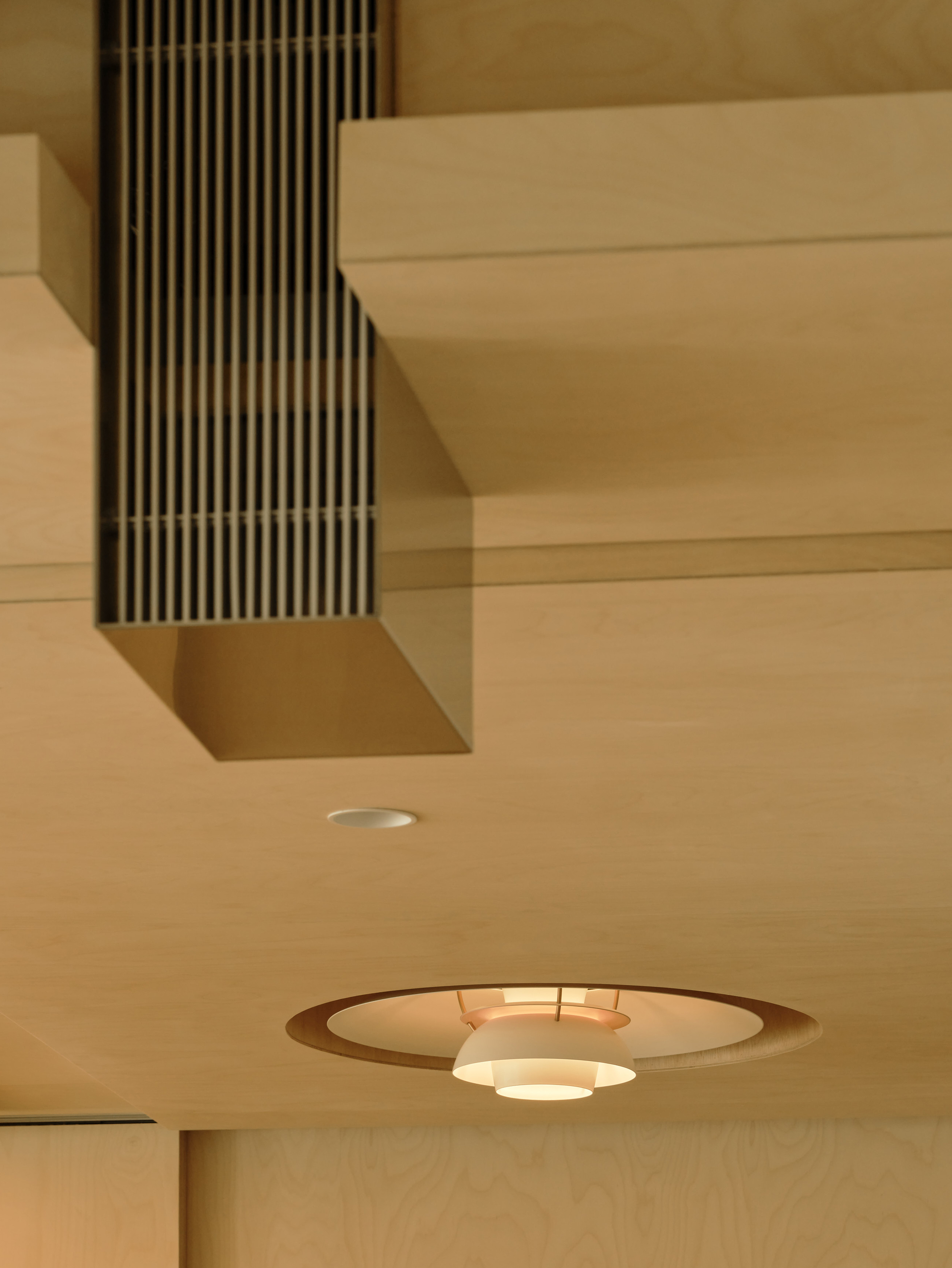 Birch plywood ceiling with a recessed circular pendant lamp and a vertical slatted vent duct, warmly lit