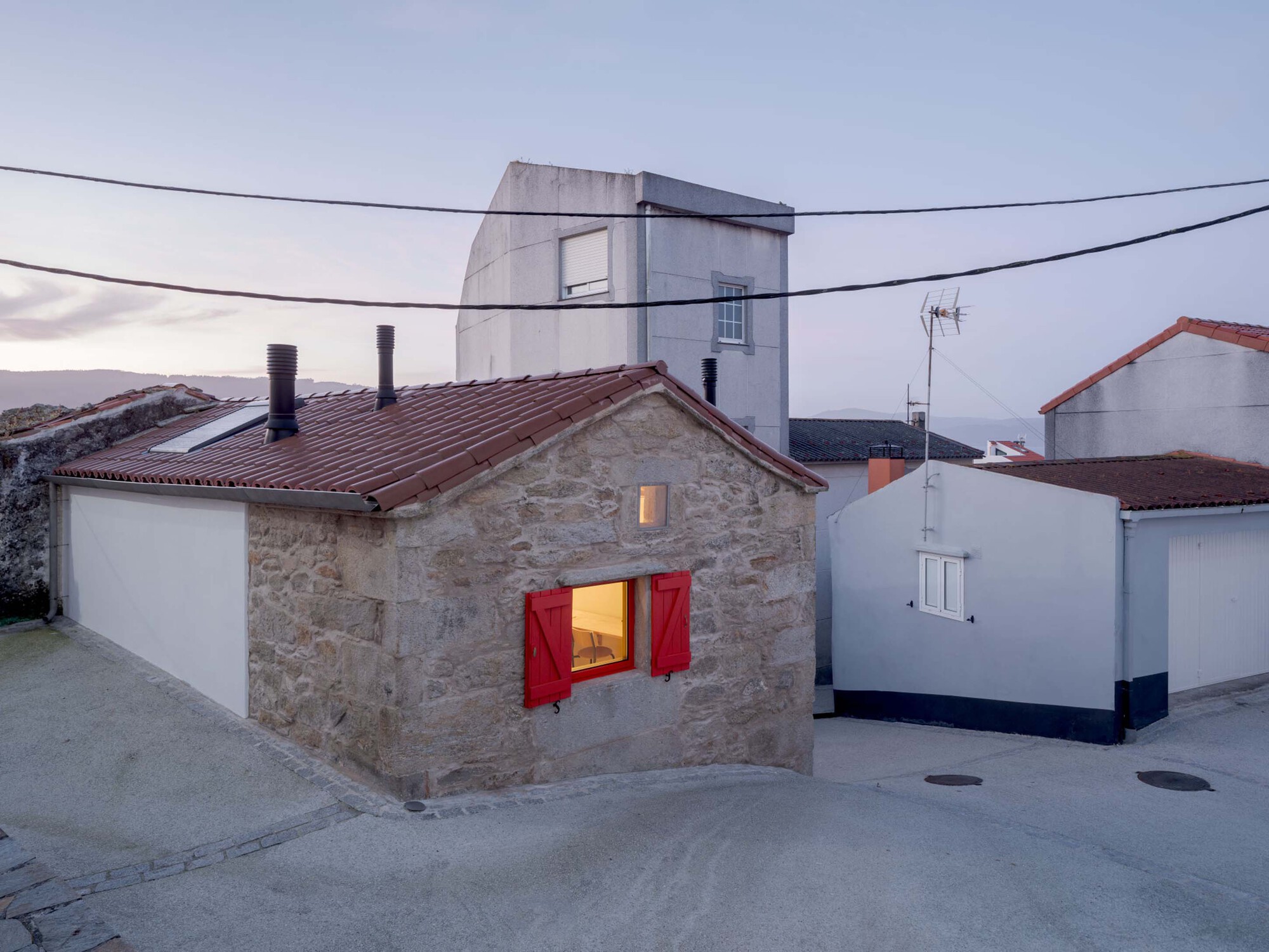 Stone gable end with open red shutters and a lit window under a red tile roof at dusk