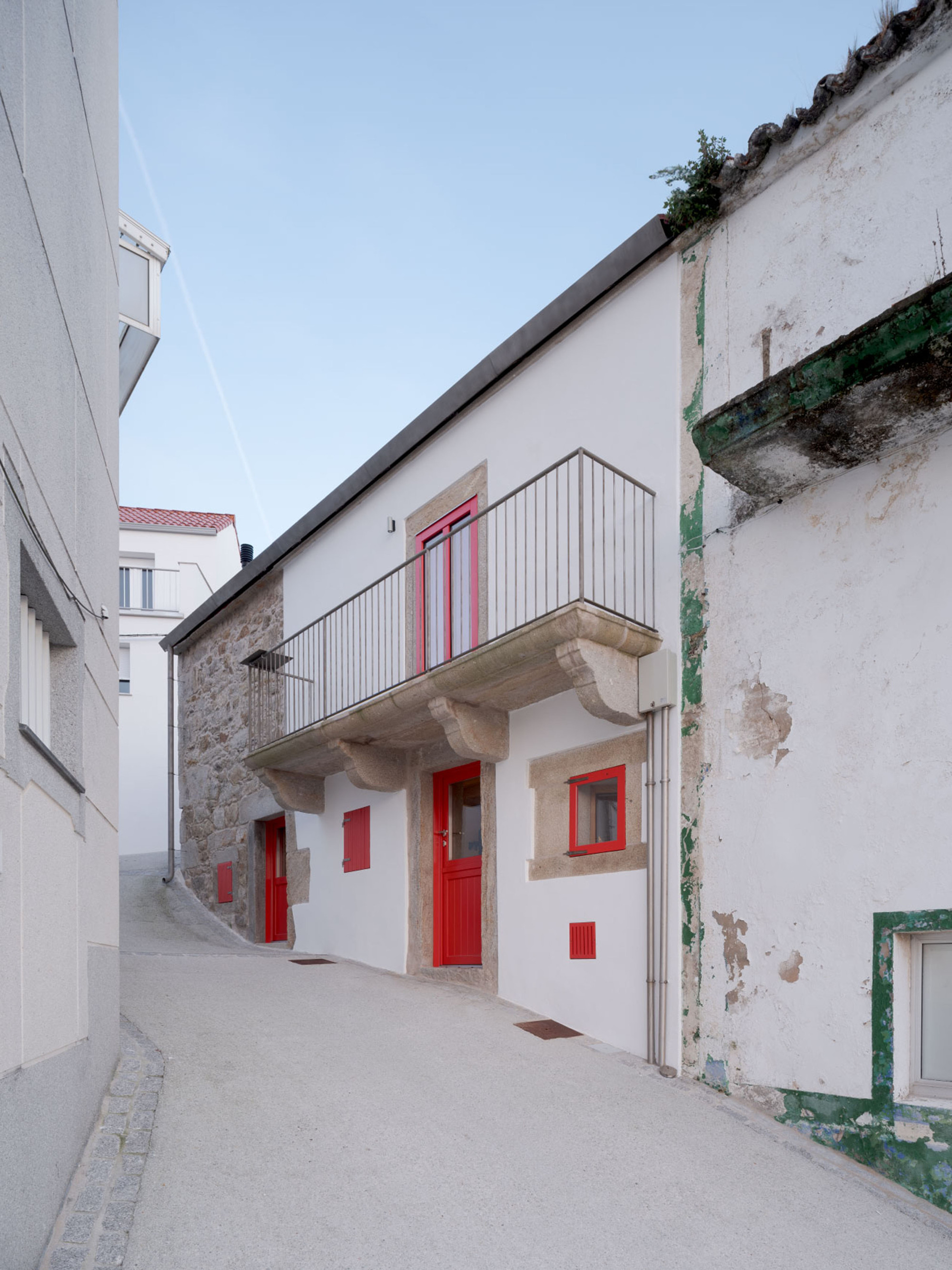 Narrow lane facing a white facade with a granite corbelled balcony and red doors under a clear blue sky