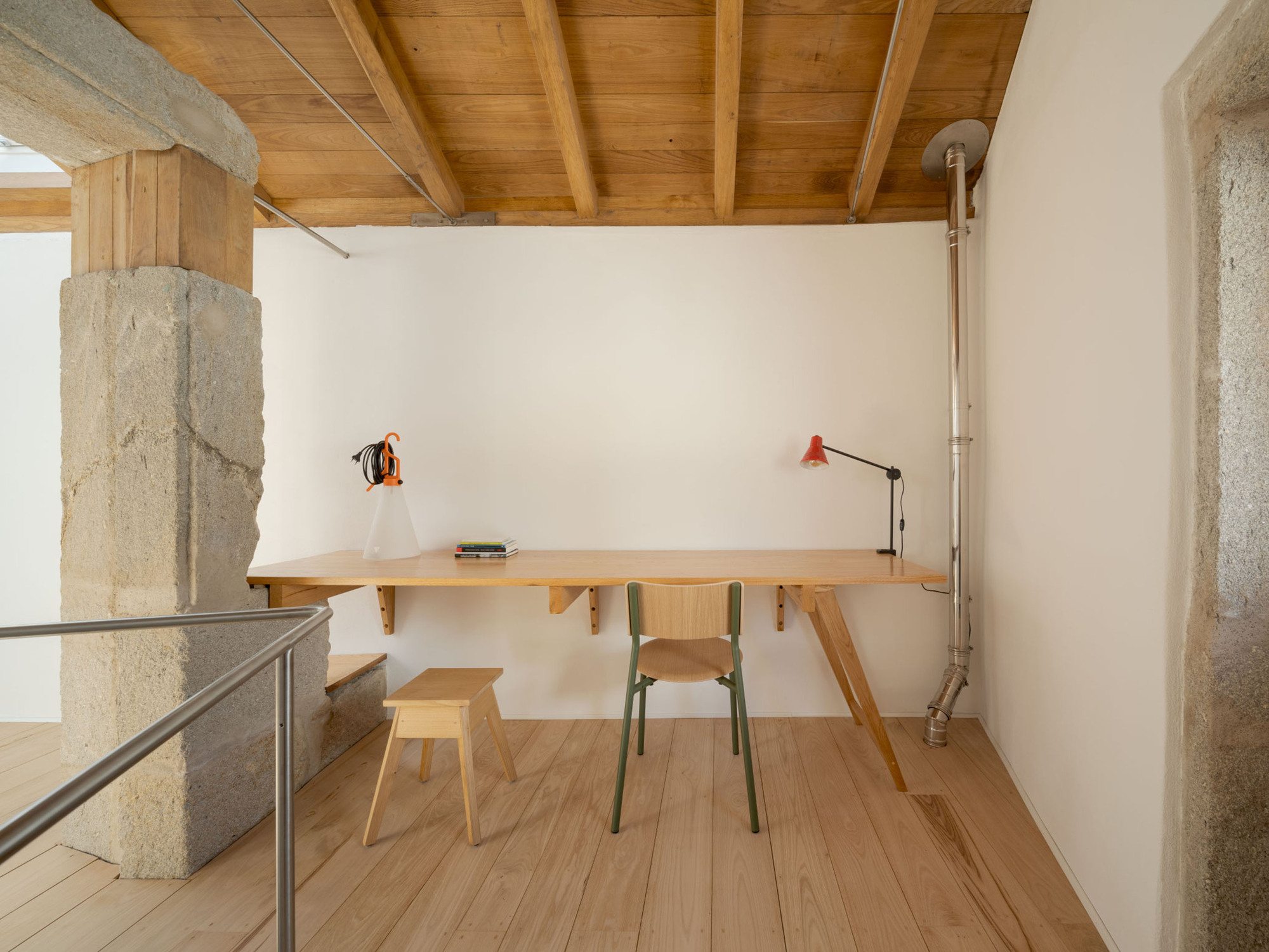 Wall-mounted timber desk with a green chair, stool and red lamp beside a rough granite pier