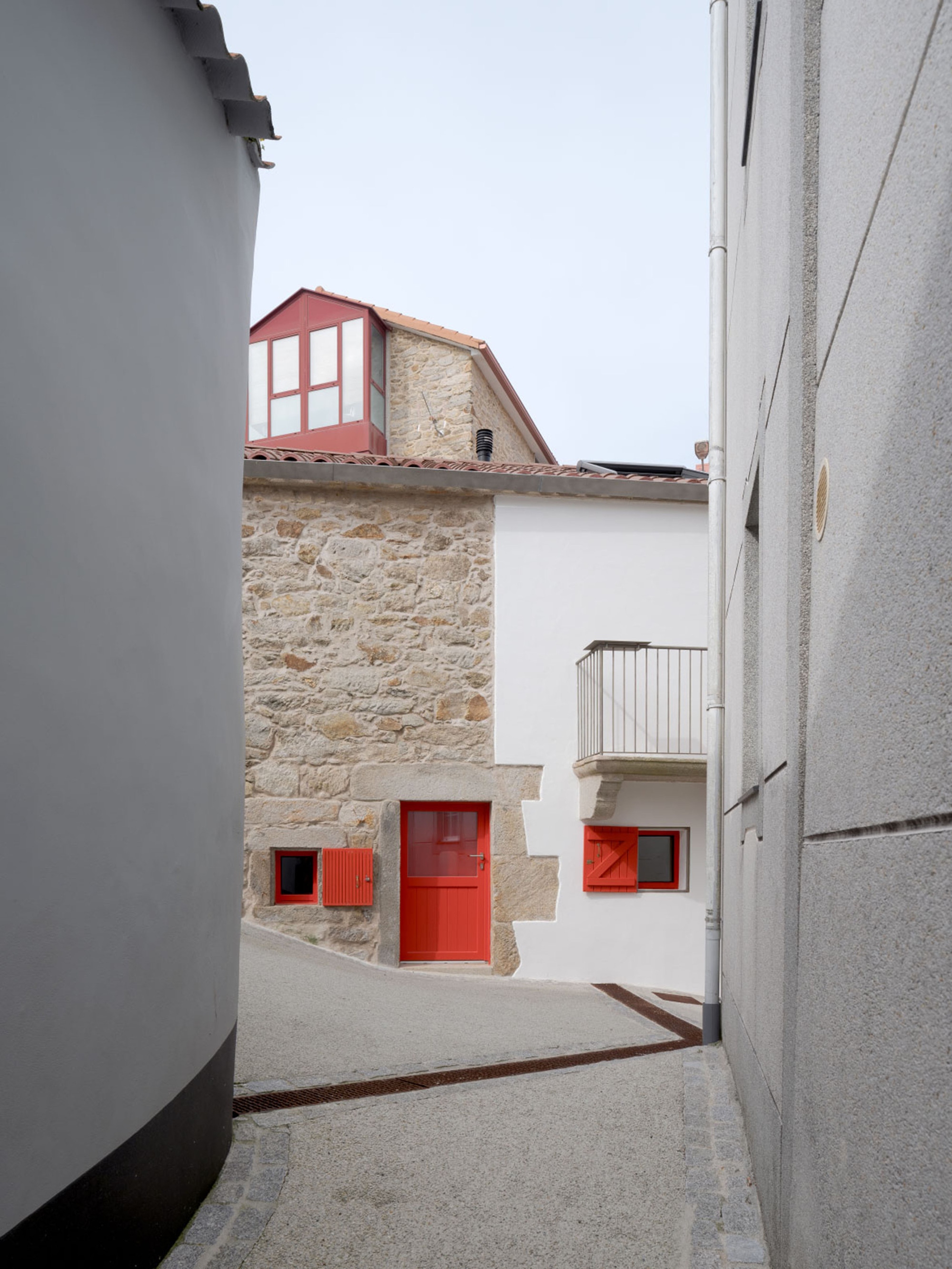 View between two grey walls toward a stone and white rendered house with red door, shutters and a red-framed dormer
