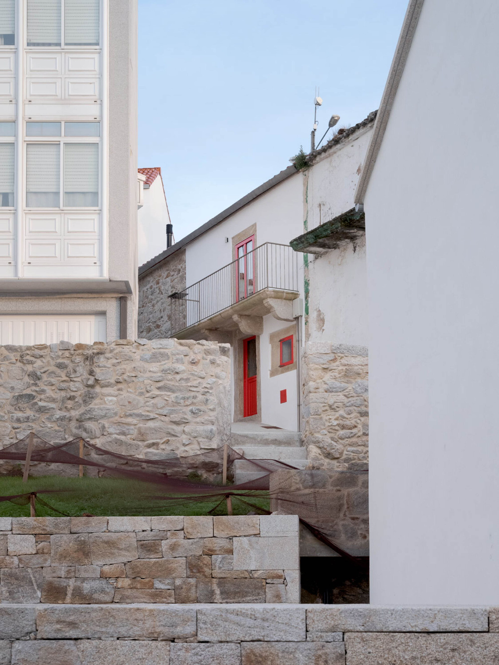 Stone village houses with red doors and a small balcony, seen between white walls and granite terraces under a pale evening sky