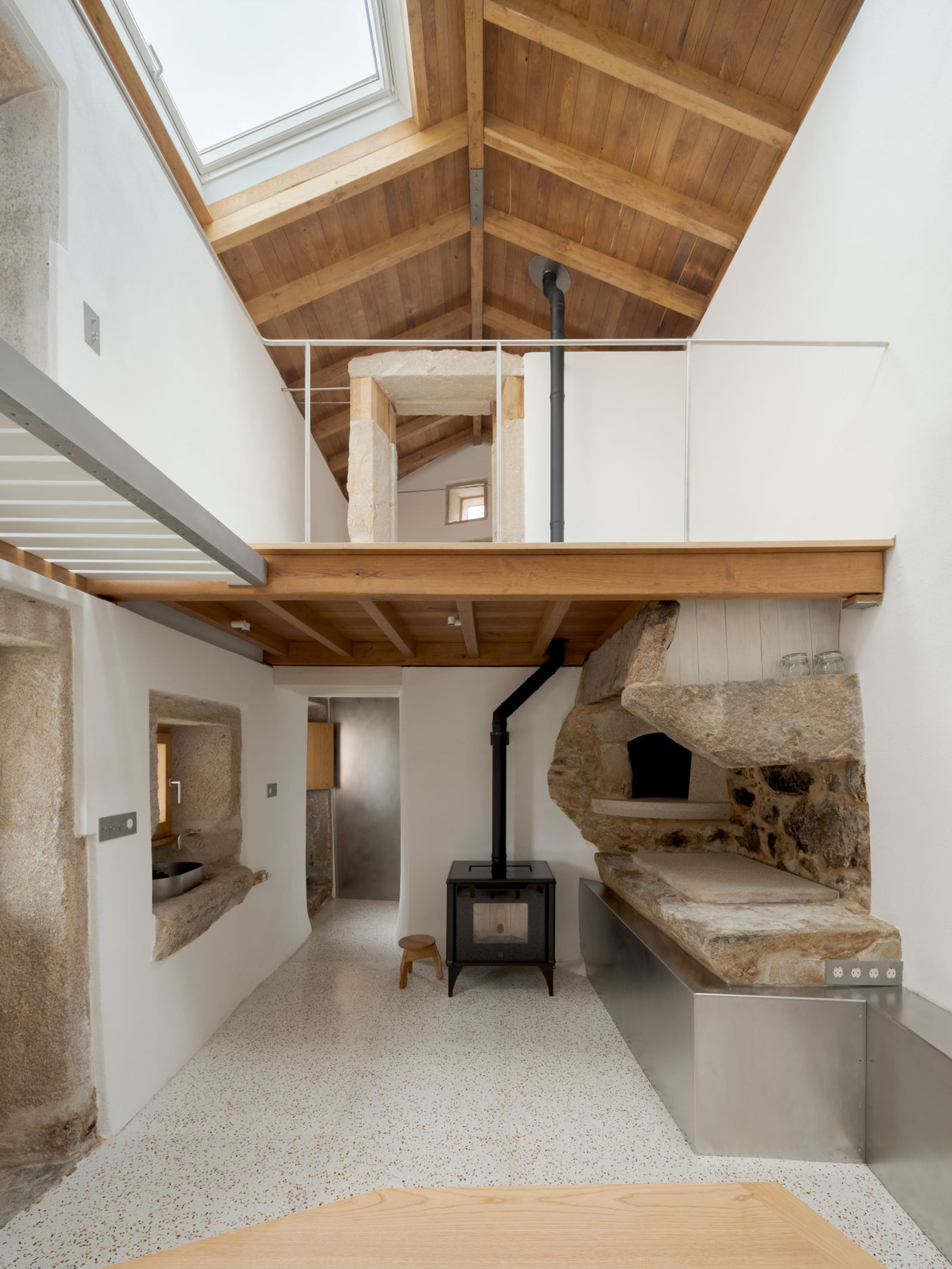 Double-height room with a black wood stove, timber mezzanine, rough granite oven niche and skylight casting daylight over the terrazzo floor