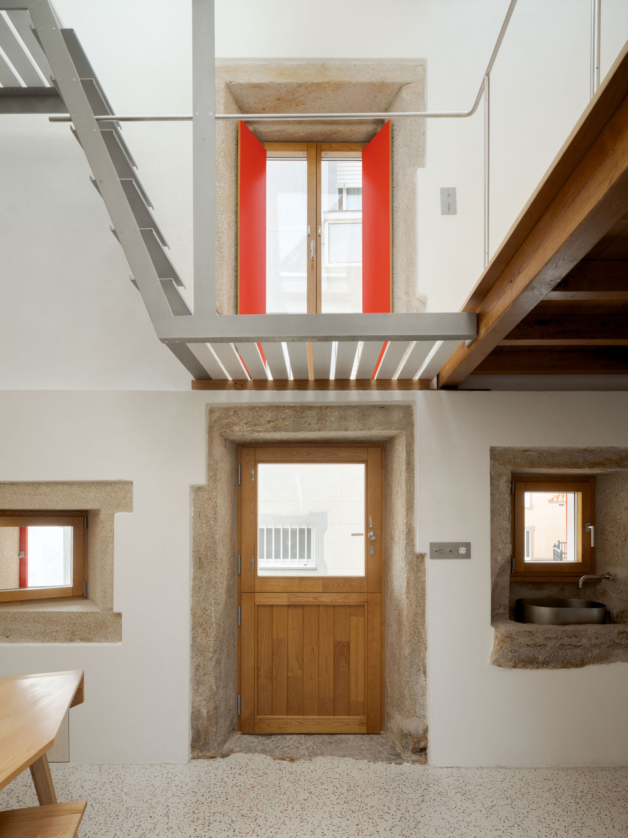 Interior wall with a timber door in a granite frame, a red-shuttered window above and a steel stair crossing overhead
