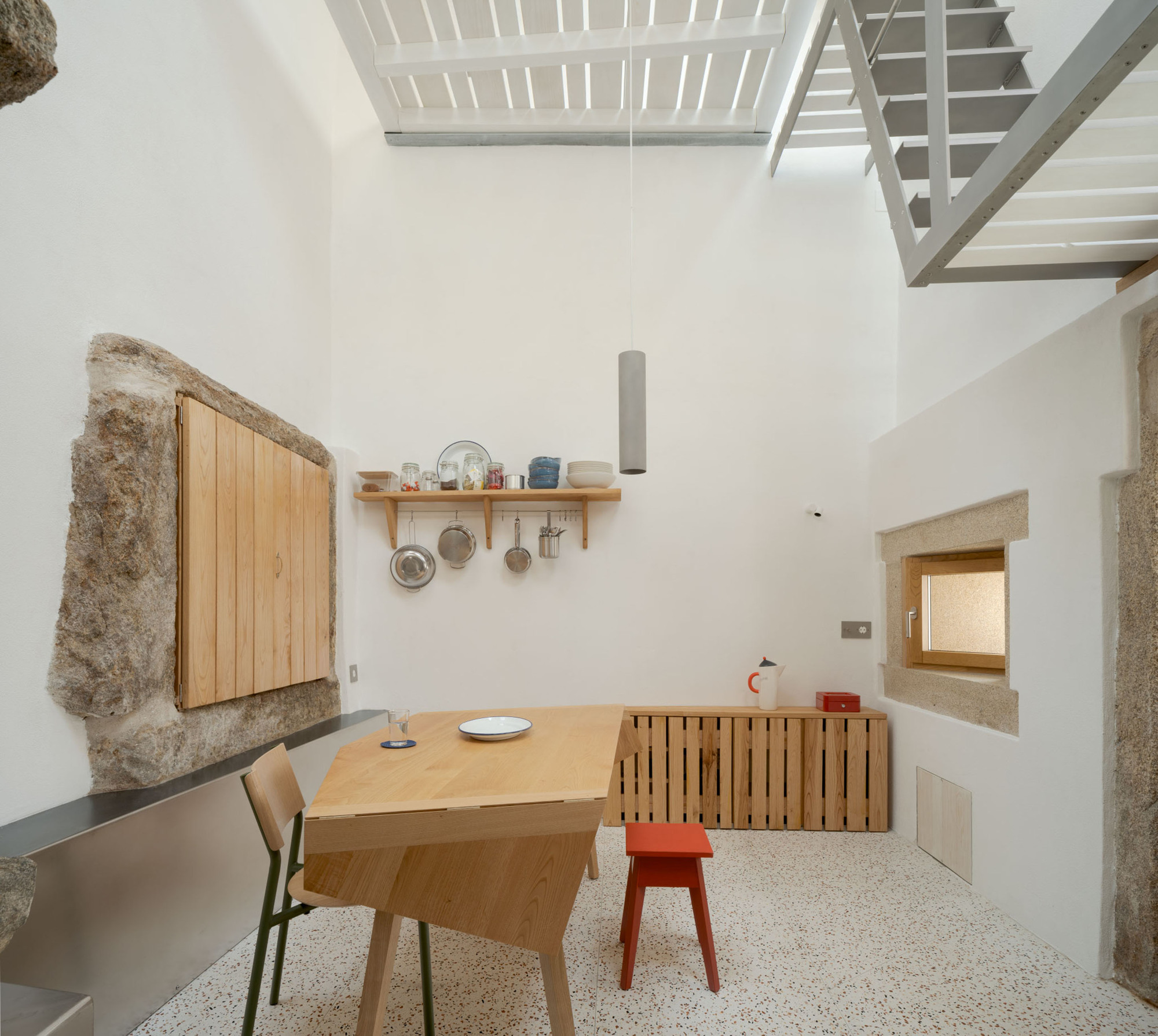 Kitchen and dining corner with a folding timber table, red stool, slatted cabinet and hanging pots, lit from a stair-side skylight