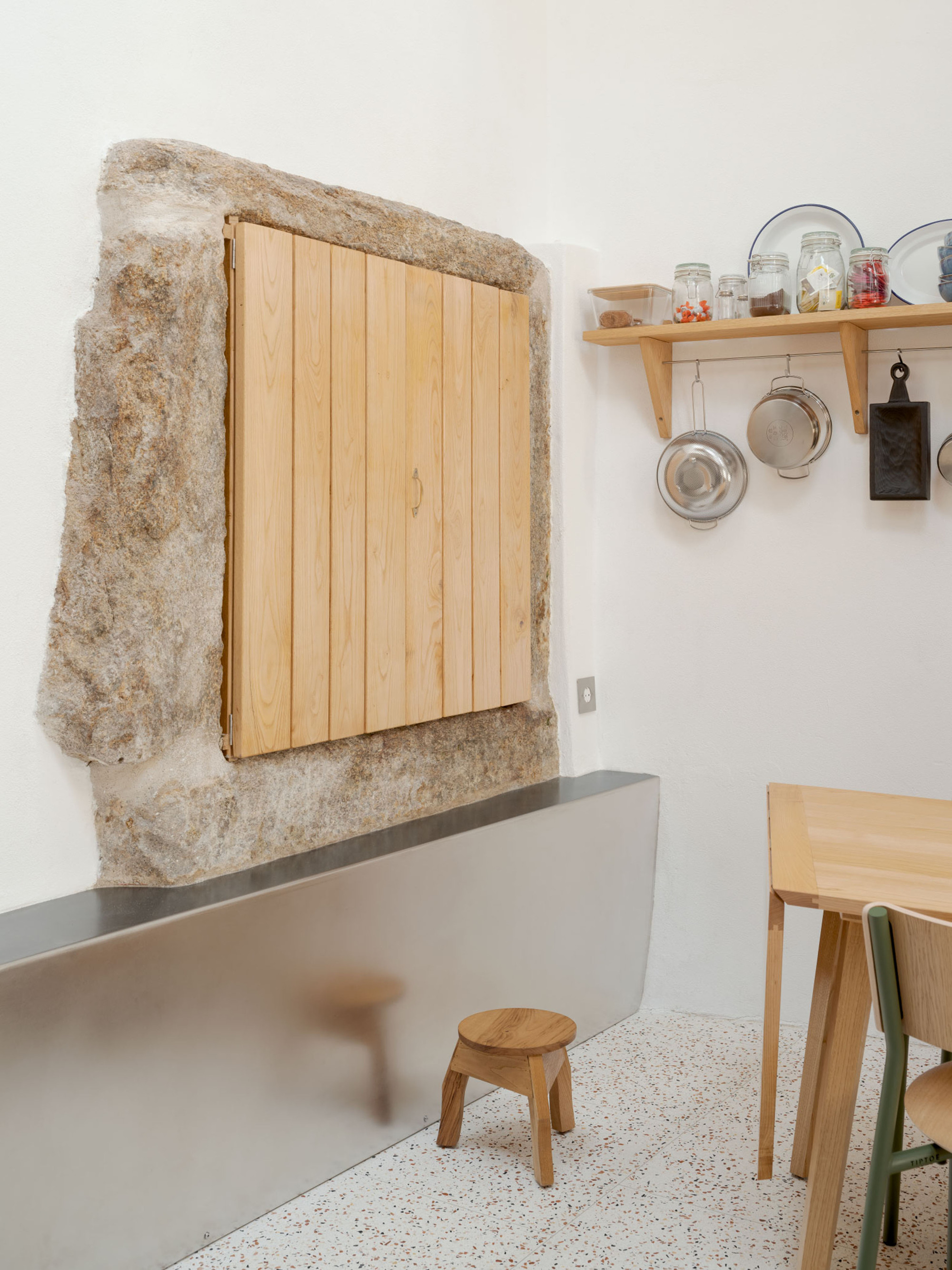 Close-up of a timber shutter set in a rough granite surround above a stainless steel counter, with a small stool and shelf of jars