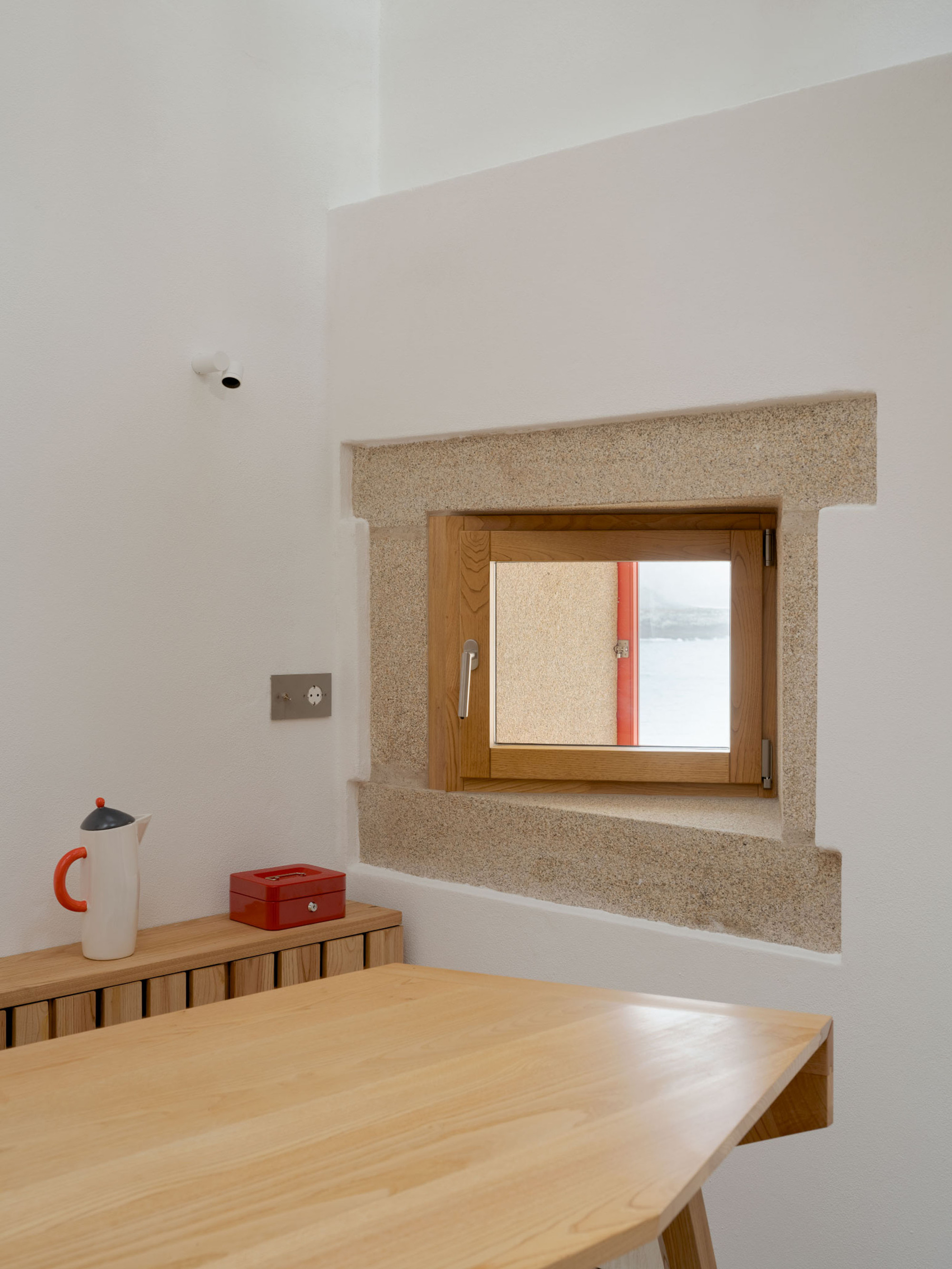 Small timber-framed window set in a granite surround above a pine table, with a red-handled jug on a slatted shelf