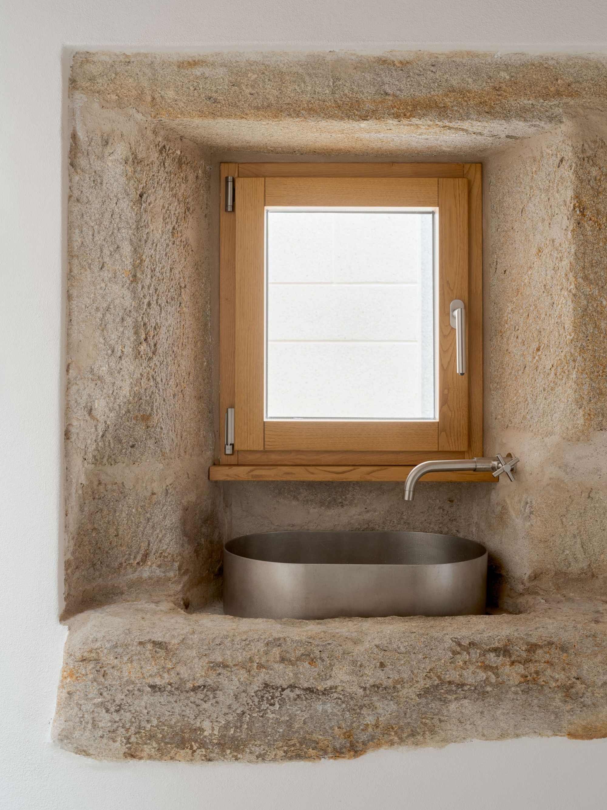 Rough granite window recess holding a timber casement above a steel basin and cross-handled tap in soft daylight