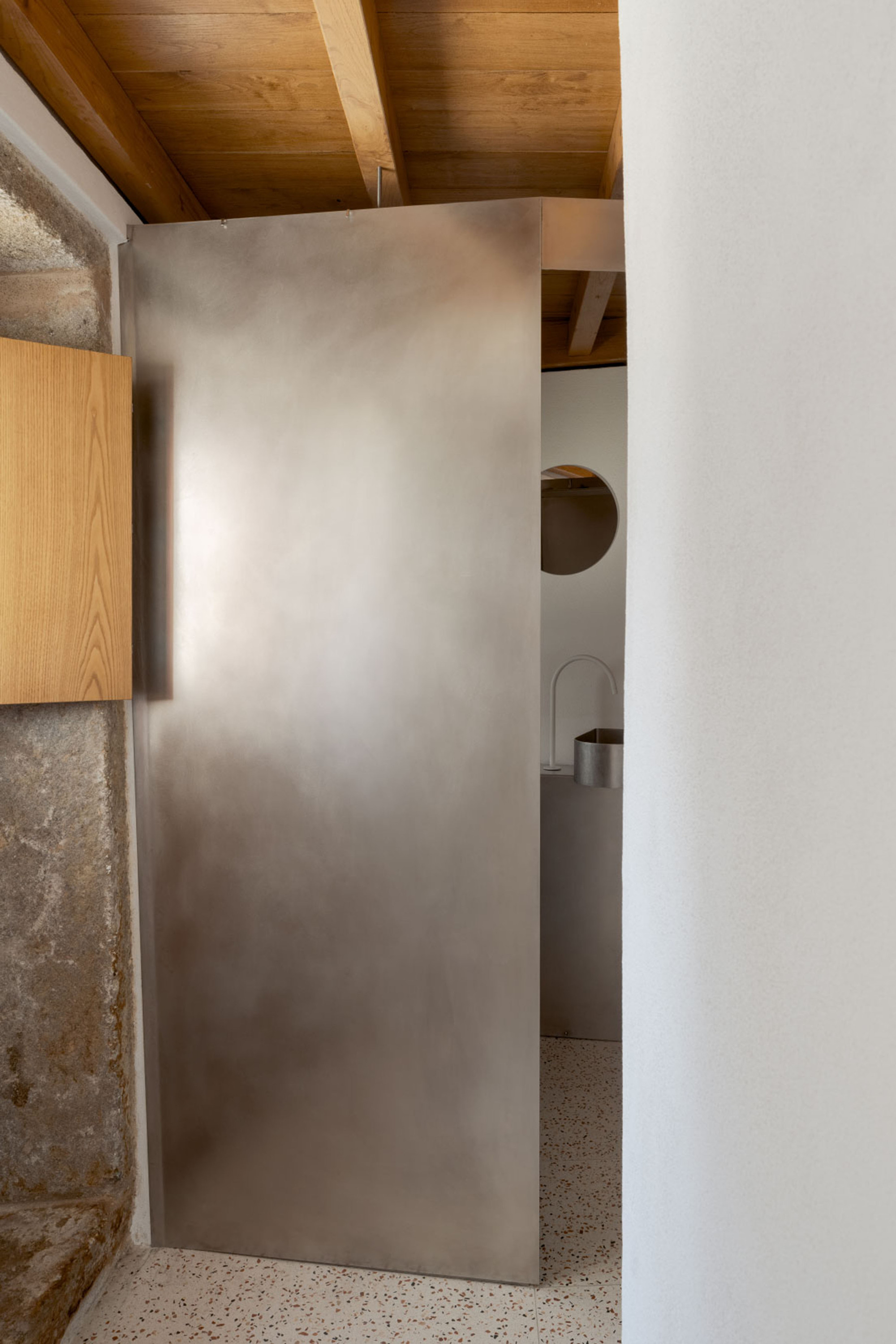 Brushed steel pivot door opening onto a terrazzo floor, with a round mirror and steel sink beyond under timber beams