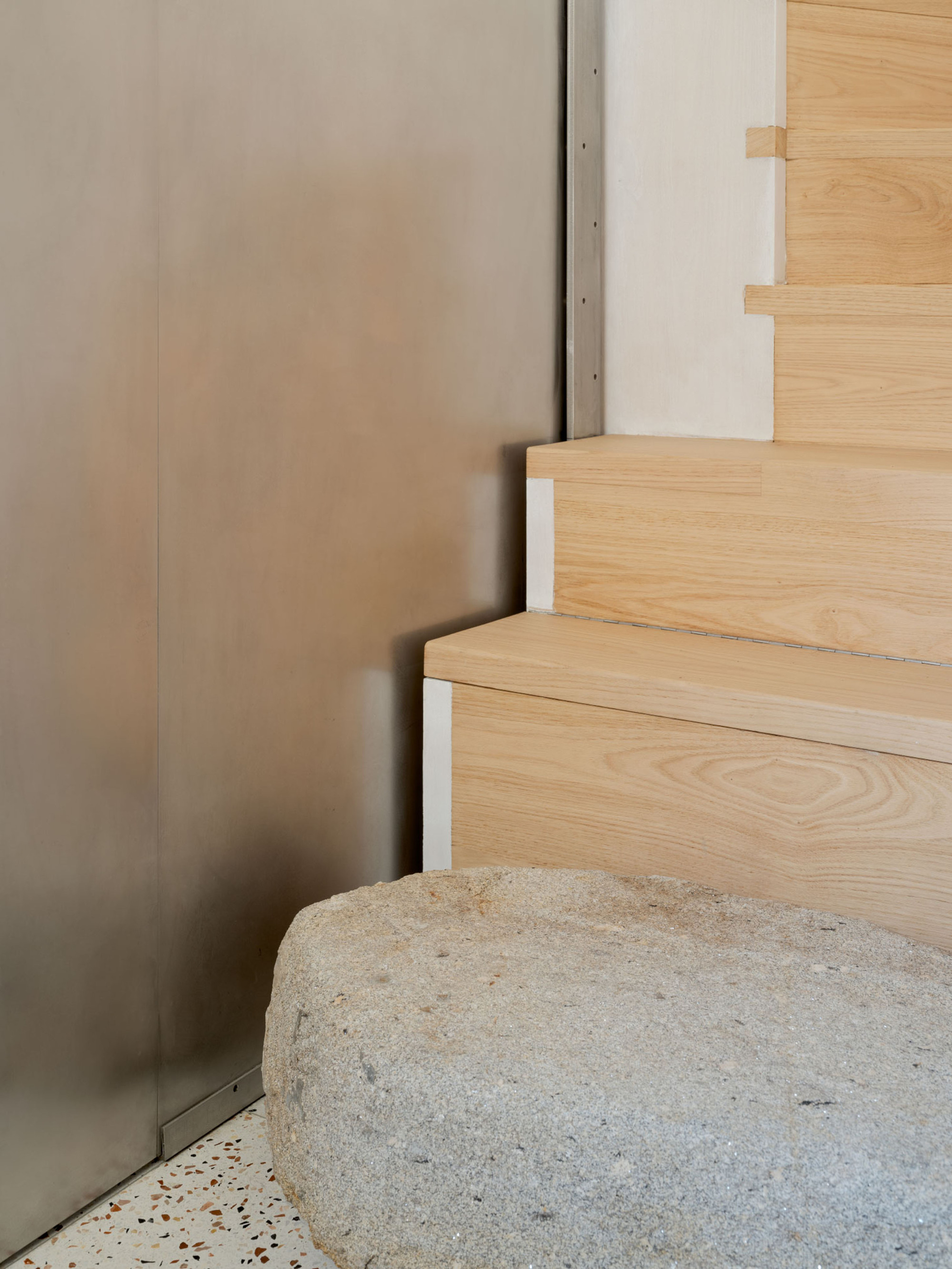 Corner of pale oak stair treads beside a brushed metal wall panel and a rough granite boulder on a terrazzo floor