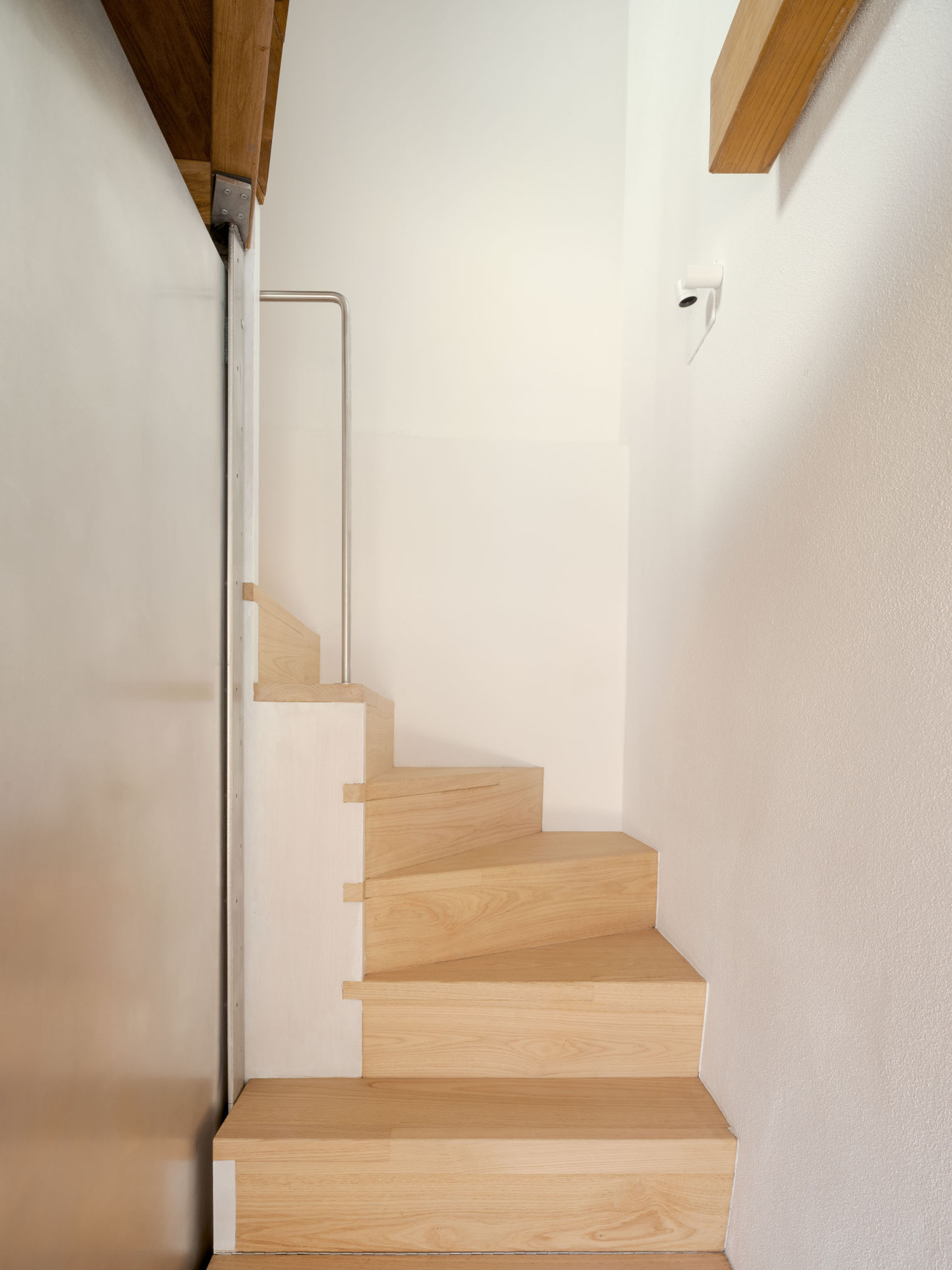 Narrow oak staircase with winding treads between a white plaster wall and a metal panel, lit by soft daylight from above