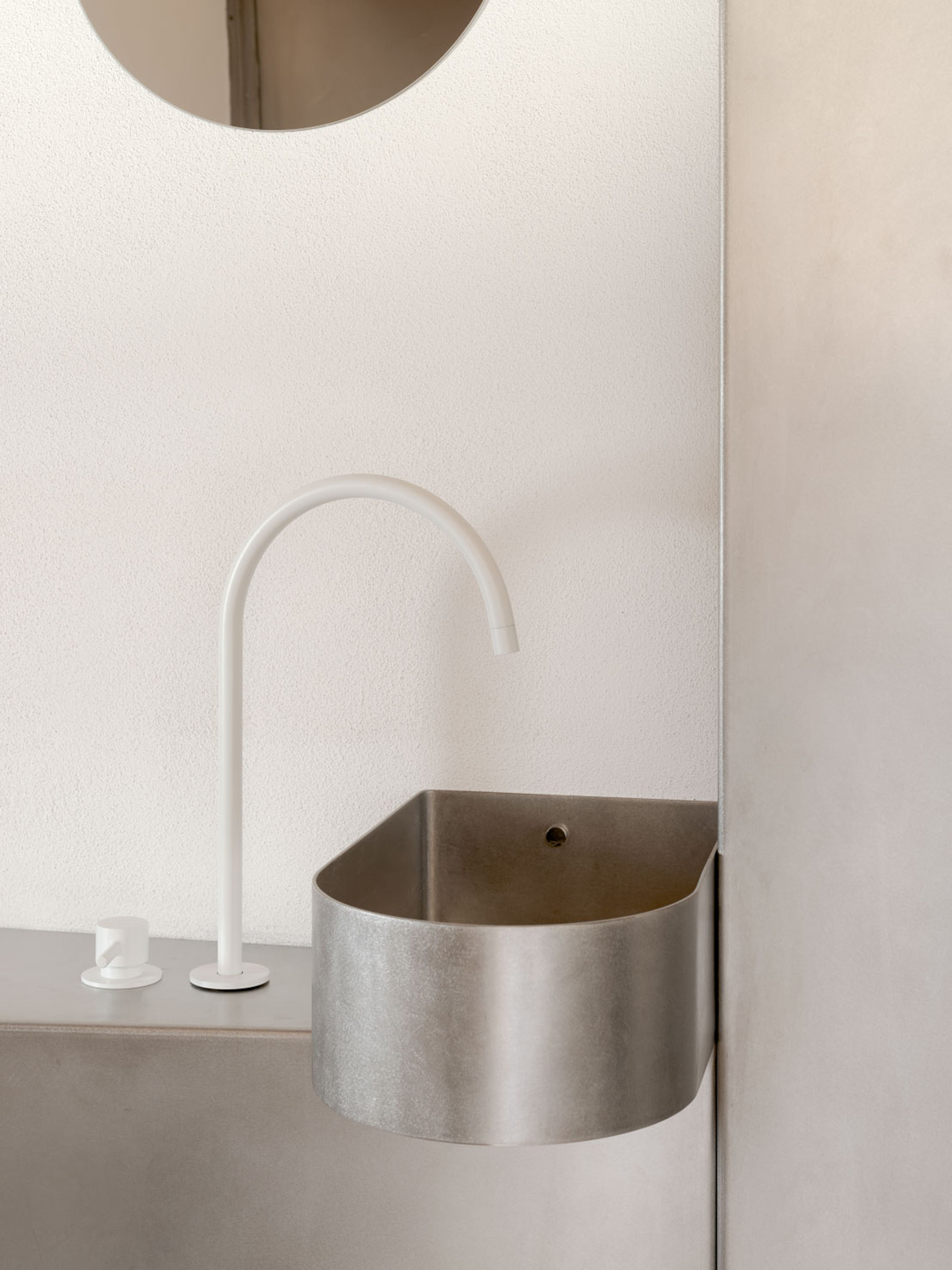 Half-round metal basin with a white arched tap on a plaster wall, a circular mirror edge above