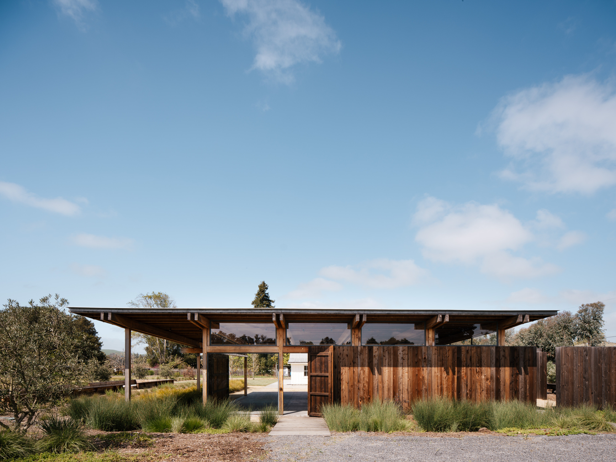 Front view of a flat-roofed timber pavilion with a vertical board screen wall and glazed clerestory under a blue sky with scattered clouds