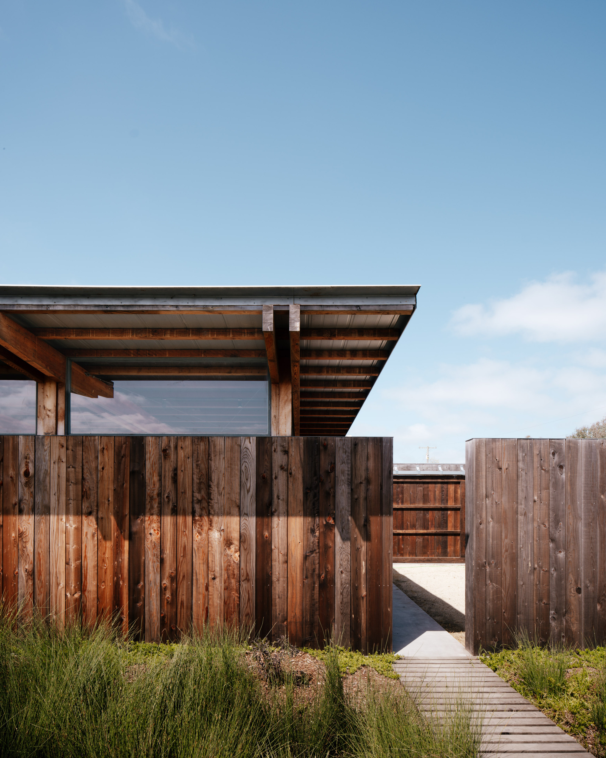 Vertical weathered board walls, a glazed band and roof overhang, with a wooden boardwalk leading through grasses toward a gap in the fence