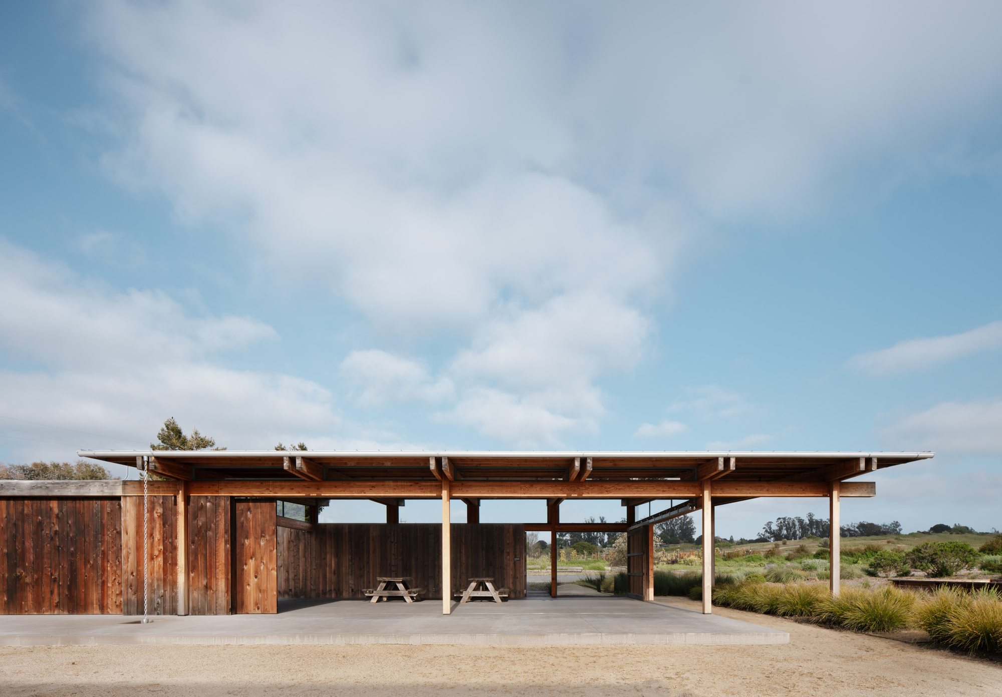 Straight-on view of a low timber pavilion with a flat overhanging roof, picnic tables and a concrete slab under scattered clouds
