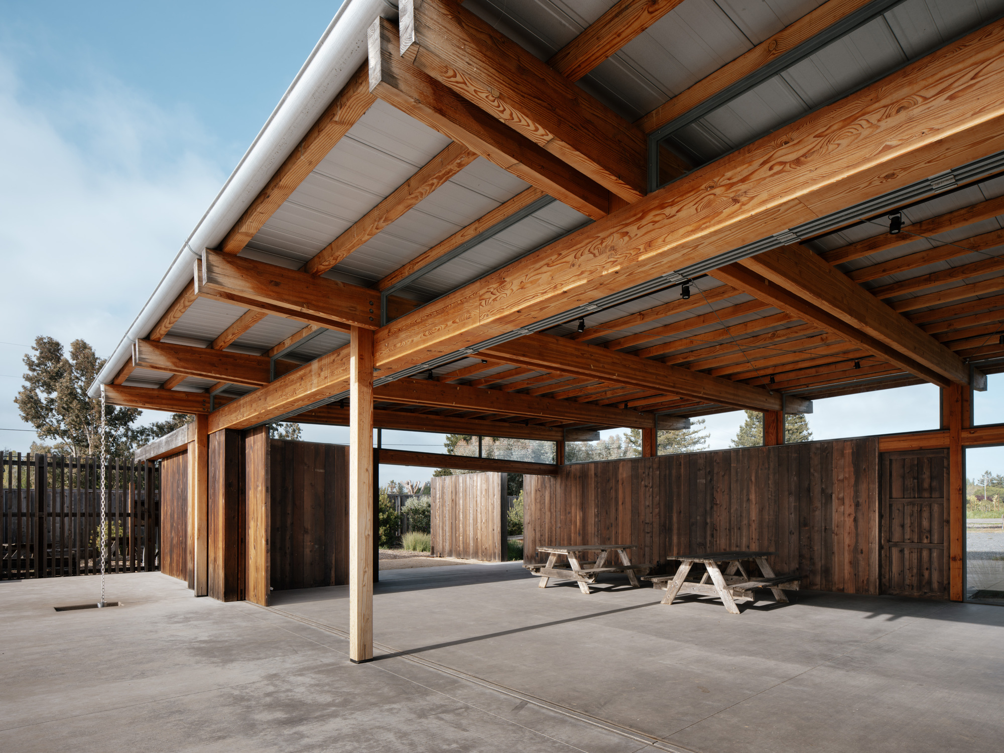 Under the roof, heavy timber beams, corrugated deck and picnic tables on a concrete floor beside weathered plank walls in daylight