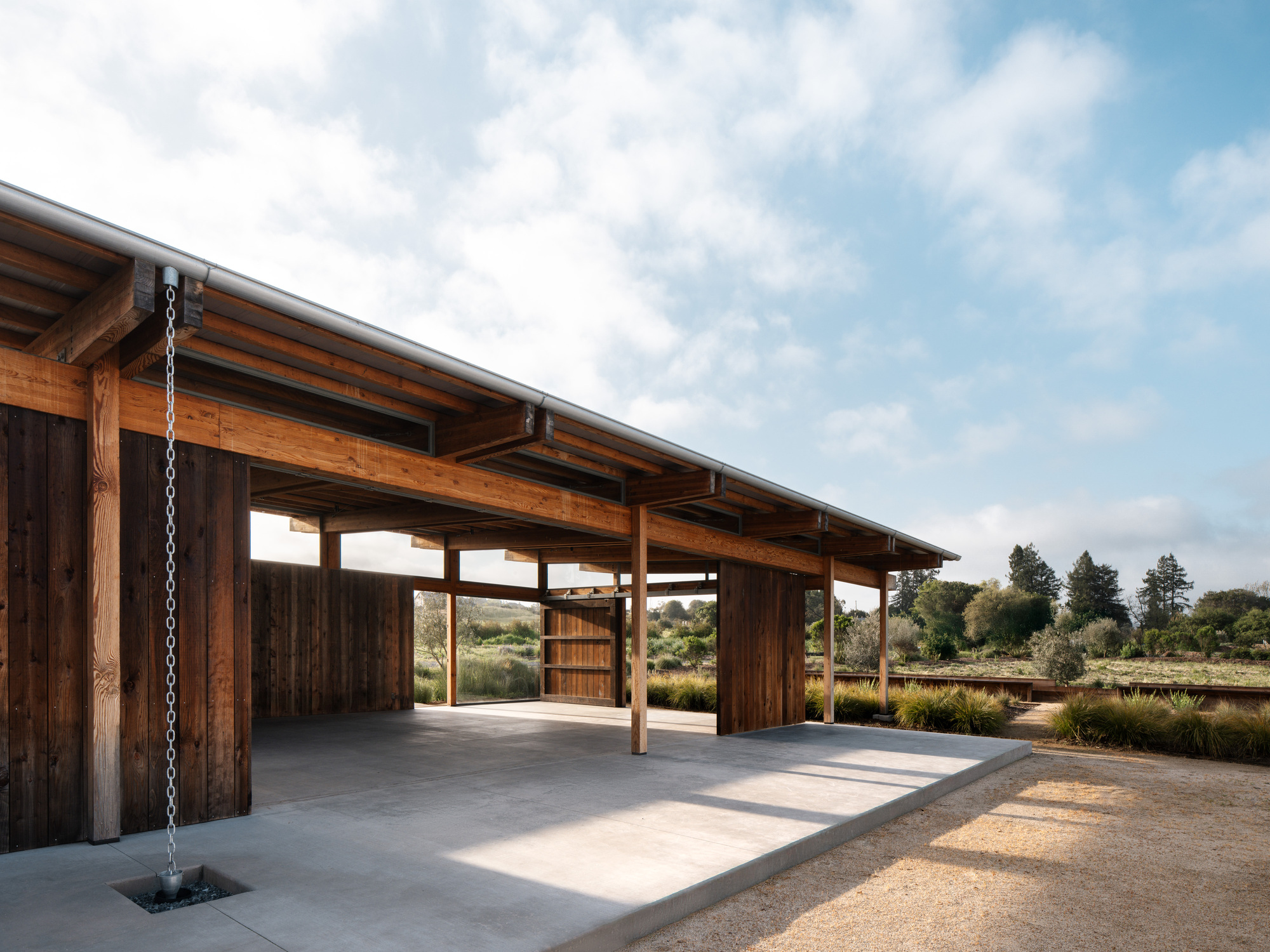 Open timber pavilion with a rain chain at the corner, concrete floor and gravel path under scattered clouds