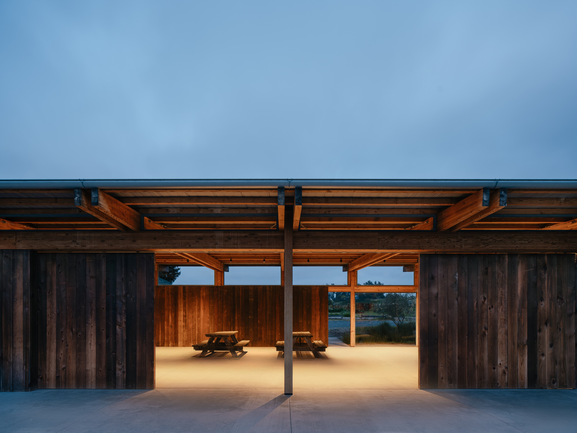 Frontal view through the open pavilion showing picnic tables on a lit floor beneath exposed beams at dusk