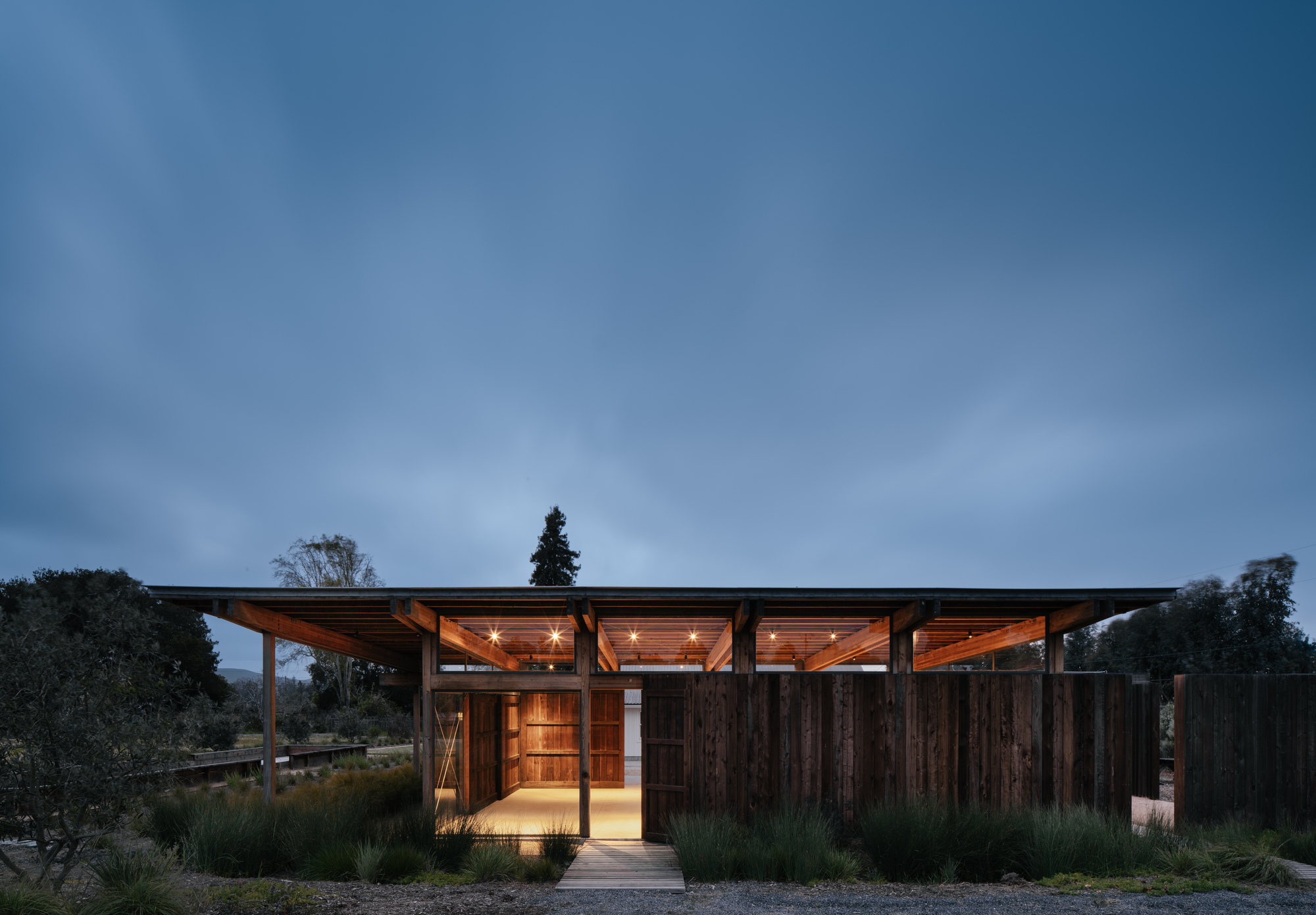 Low timber pavilion with a glowing ceiling and board-formed wood walls, surrounded by grasses under a heavy dusk sky