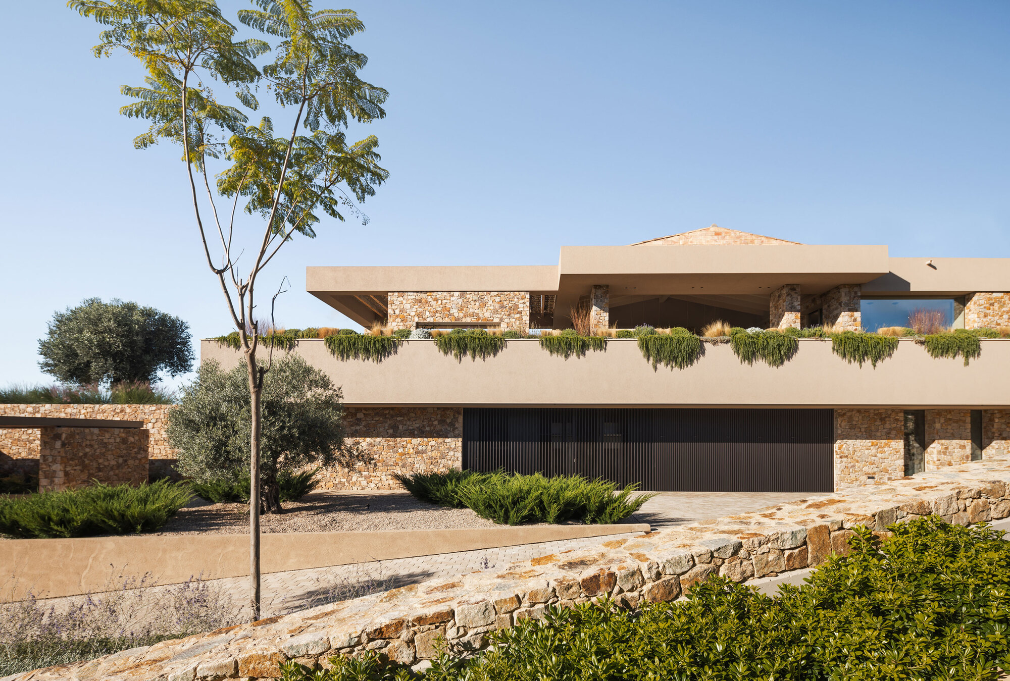 Stone and stucco house with a planted green roof edge and dark slatted garage door under a clear blue sky