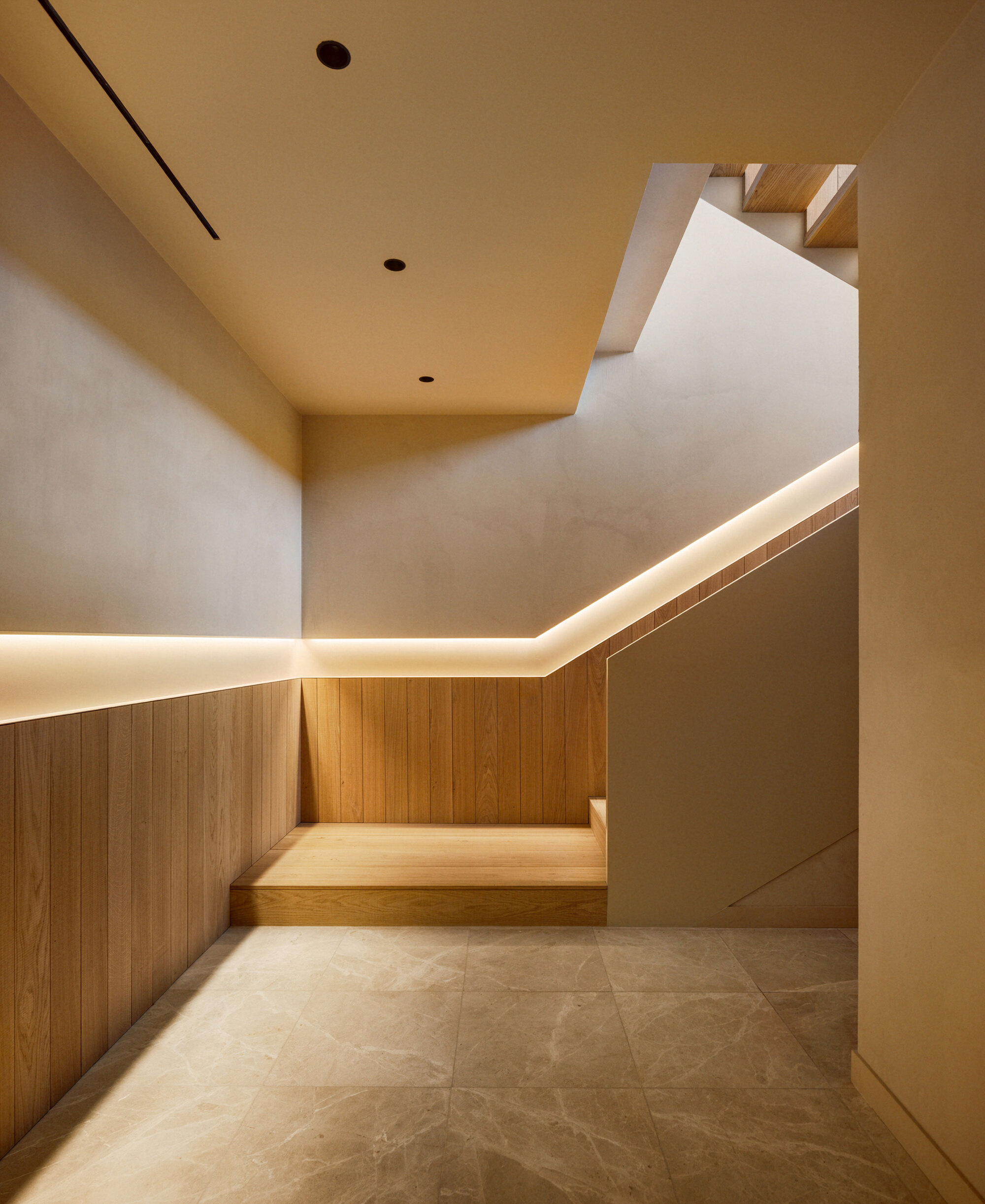 Stair hall with oak wall paneling, a continuous strip of warm light along the handrail line and a stone tile floor