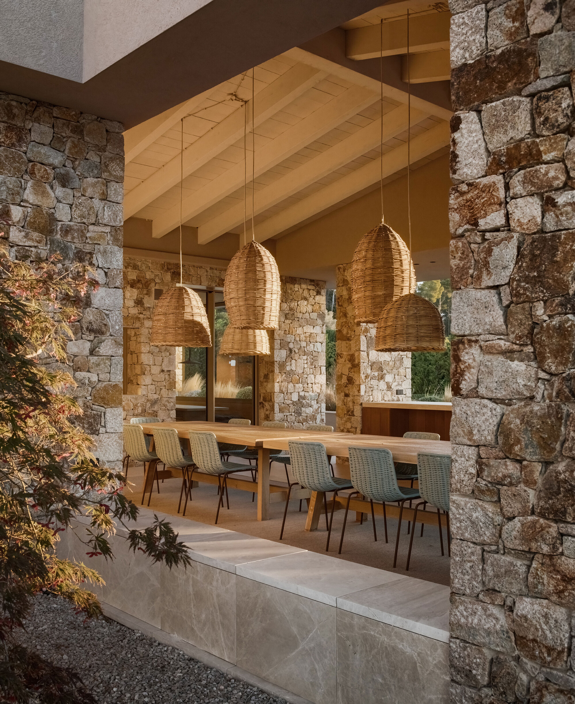 Covered dining terrace seen between stone piers, with woven pendant lamps hanging over a long timber table and exposed rafters