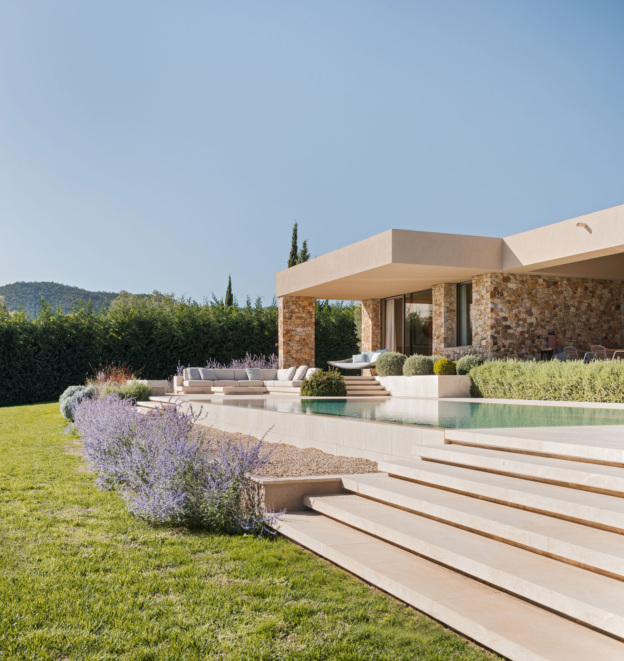 Wide limestone steps leading to an infinity pool and a stone-columned pavilion, with lavender in the foreground lawn under a clear sky