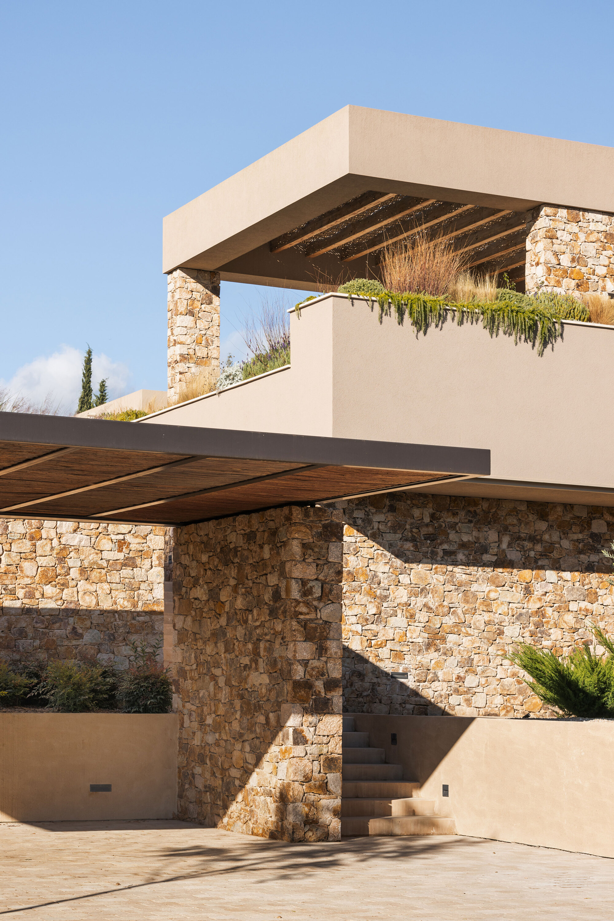 Stone pier and timber-slatted canopy beneath a planted rooftop terrace with grasses, under a clear blue sky