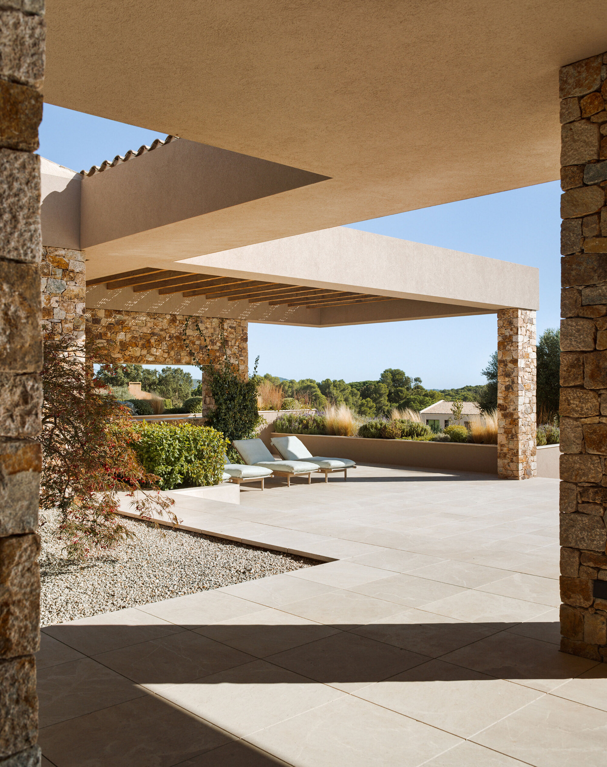Covered terrace with stone columns, pale paving and two lounge chairs facing a gravel bed and wooded hills in sunlight