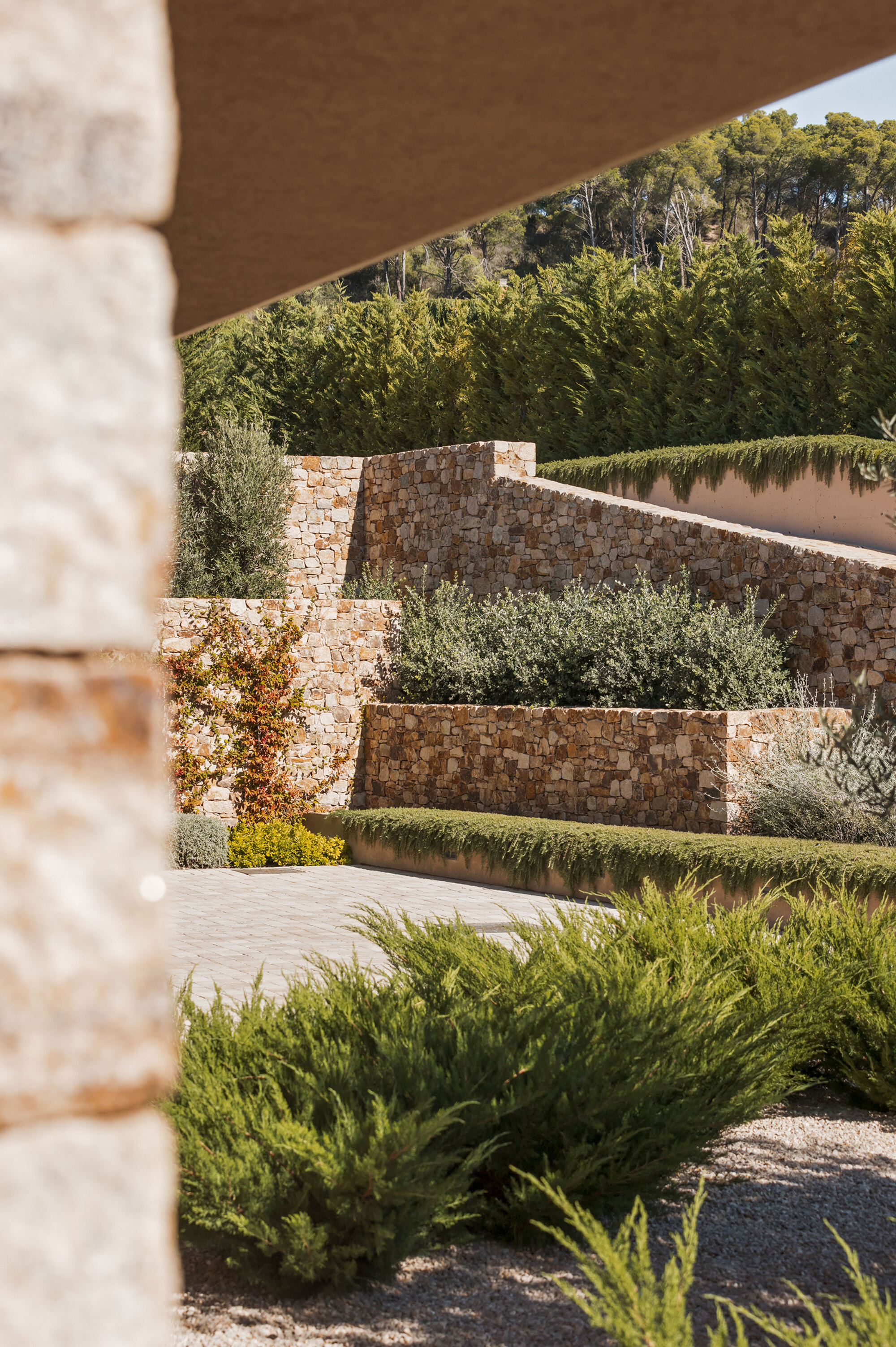 Terraced dry-stone retaining walls with clipped shrubs and juniper, framed by a stone column and a tall cypress hedge