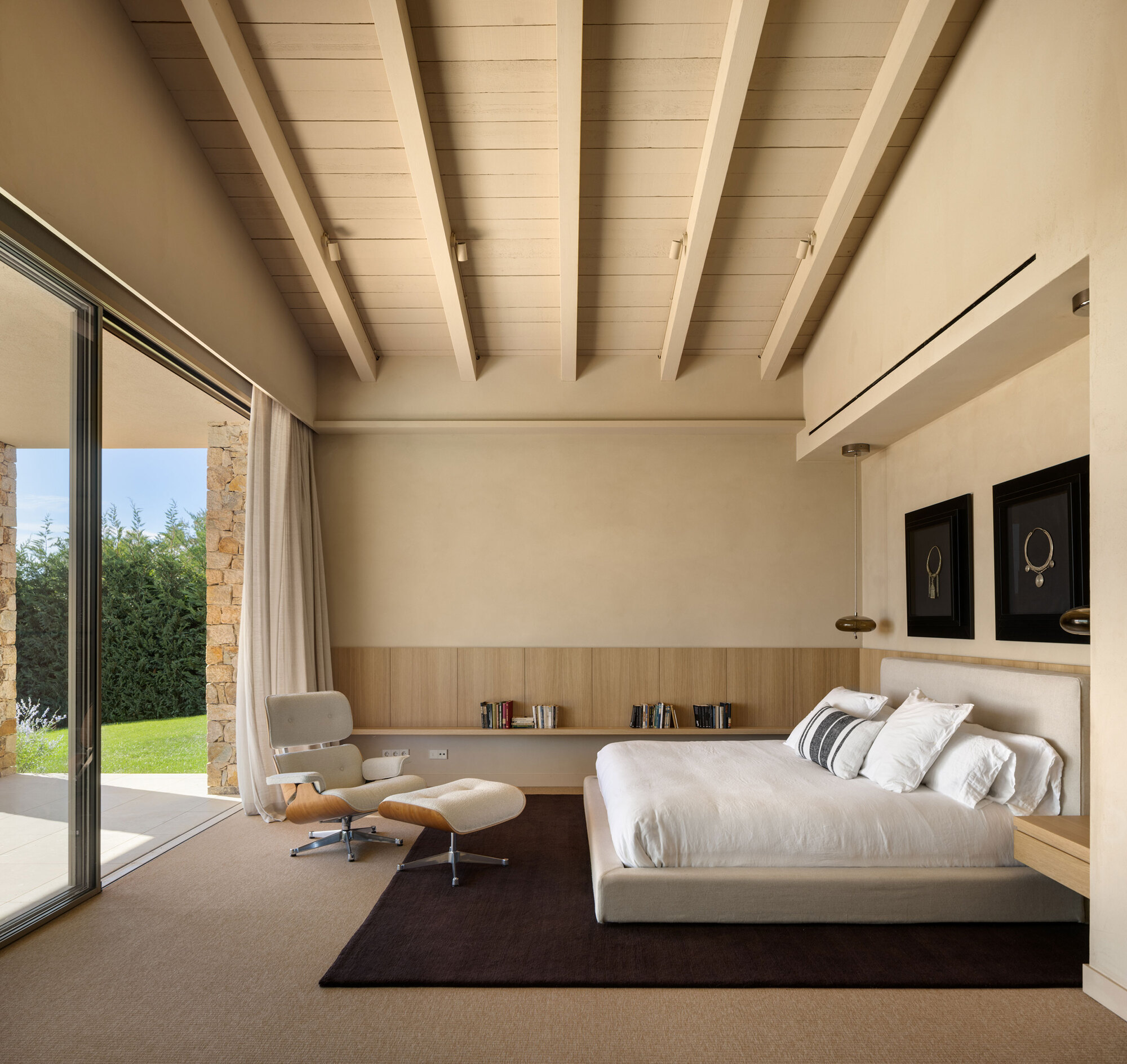 Bedroom with exposed timber beams, low wood-panelled wall, white bed and lounge chair beside a sliding glass door to the lawn
