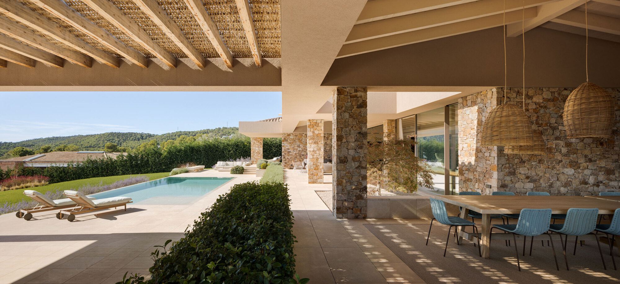 Long covered porch with stone columns, woven pendant lamps and blue chairs at a table, looking toward the pool and lawn