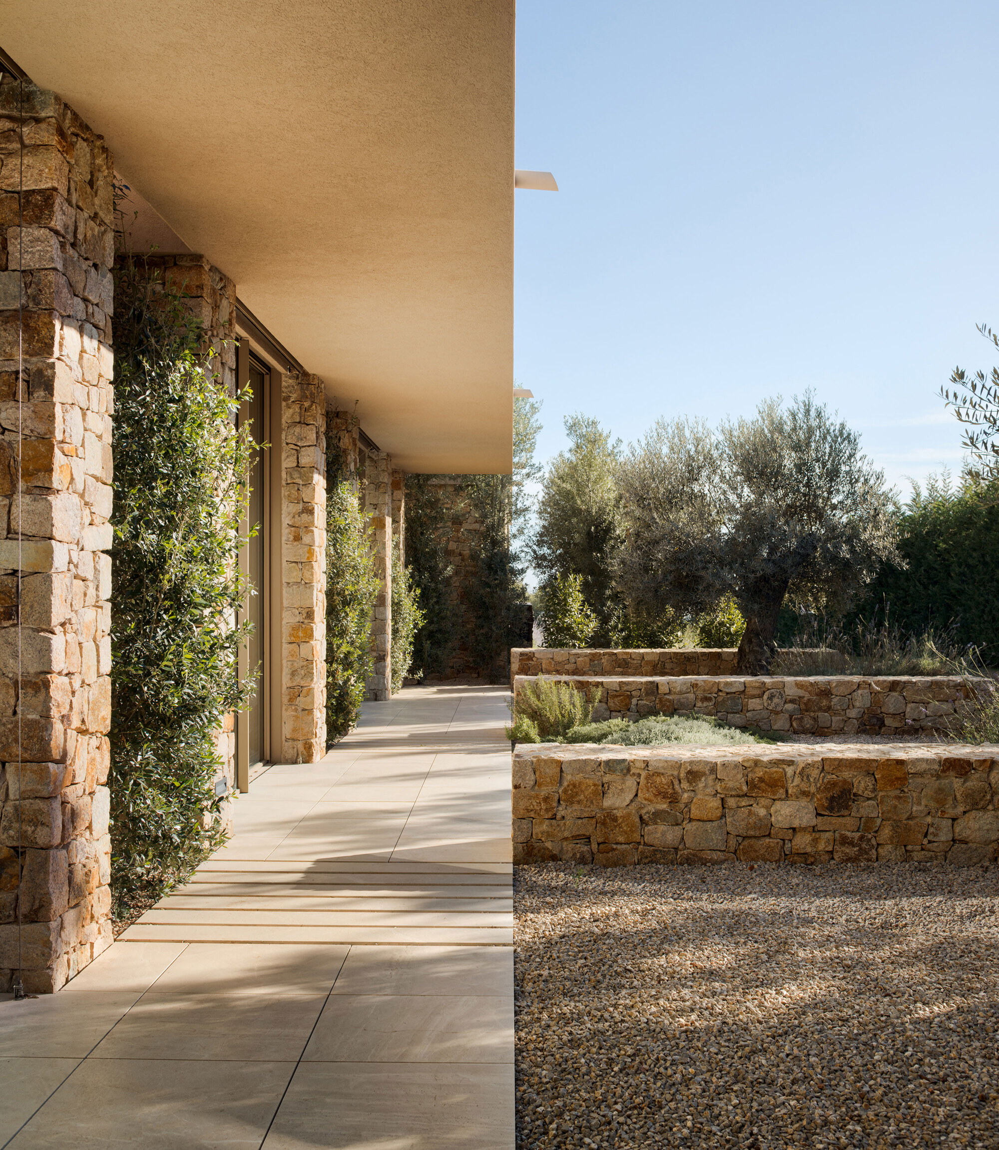 Stone-clad walkway beside a deep eave, with low stone walls, a gravel bed and an olive tree in afternoon light