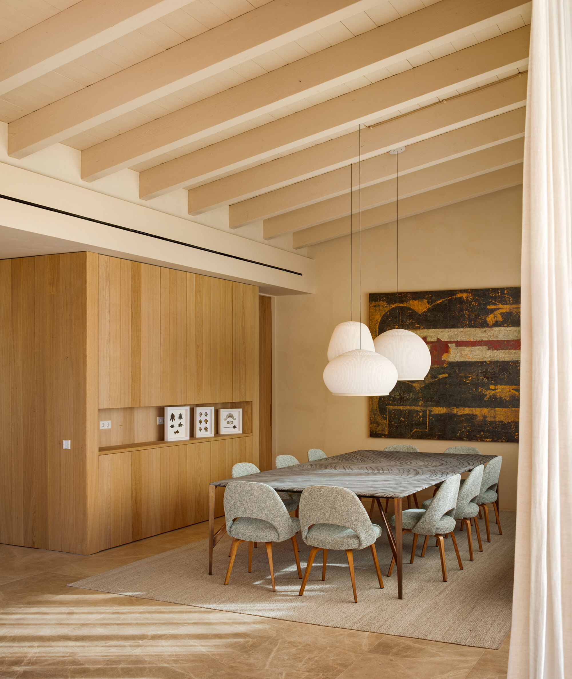 Dining room with a long stone-topped table, pale upholstered chairs, white pendant lamps and exposed whitewashed beams, with sunlight striping the floor