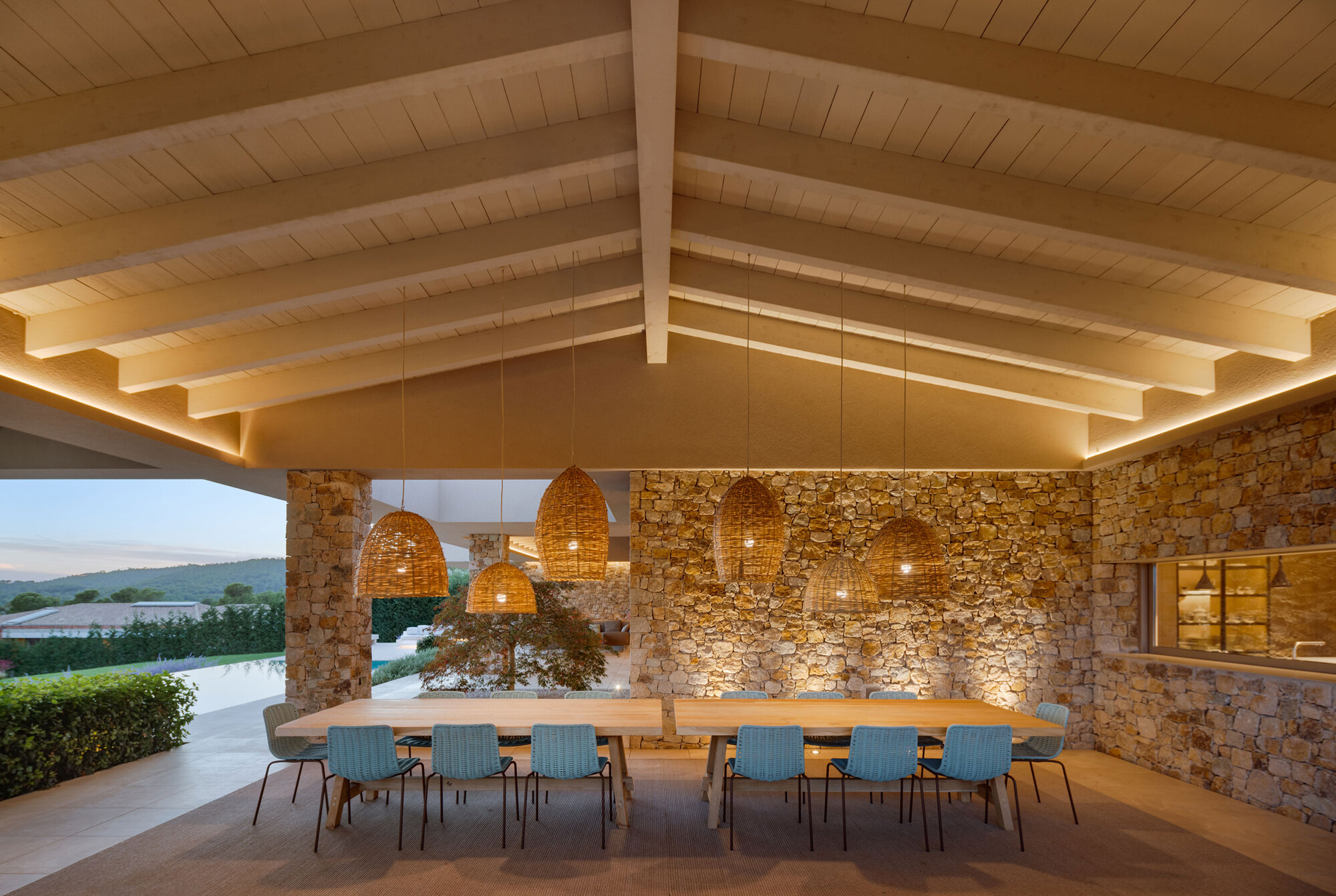 Covered terrace with a long timber table, woven pendant lamps and a rubble stone wall lit at dusk