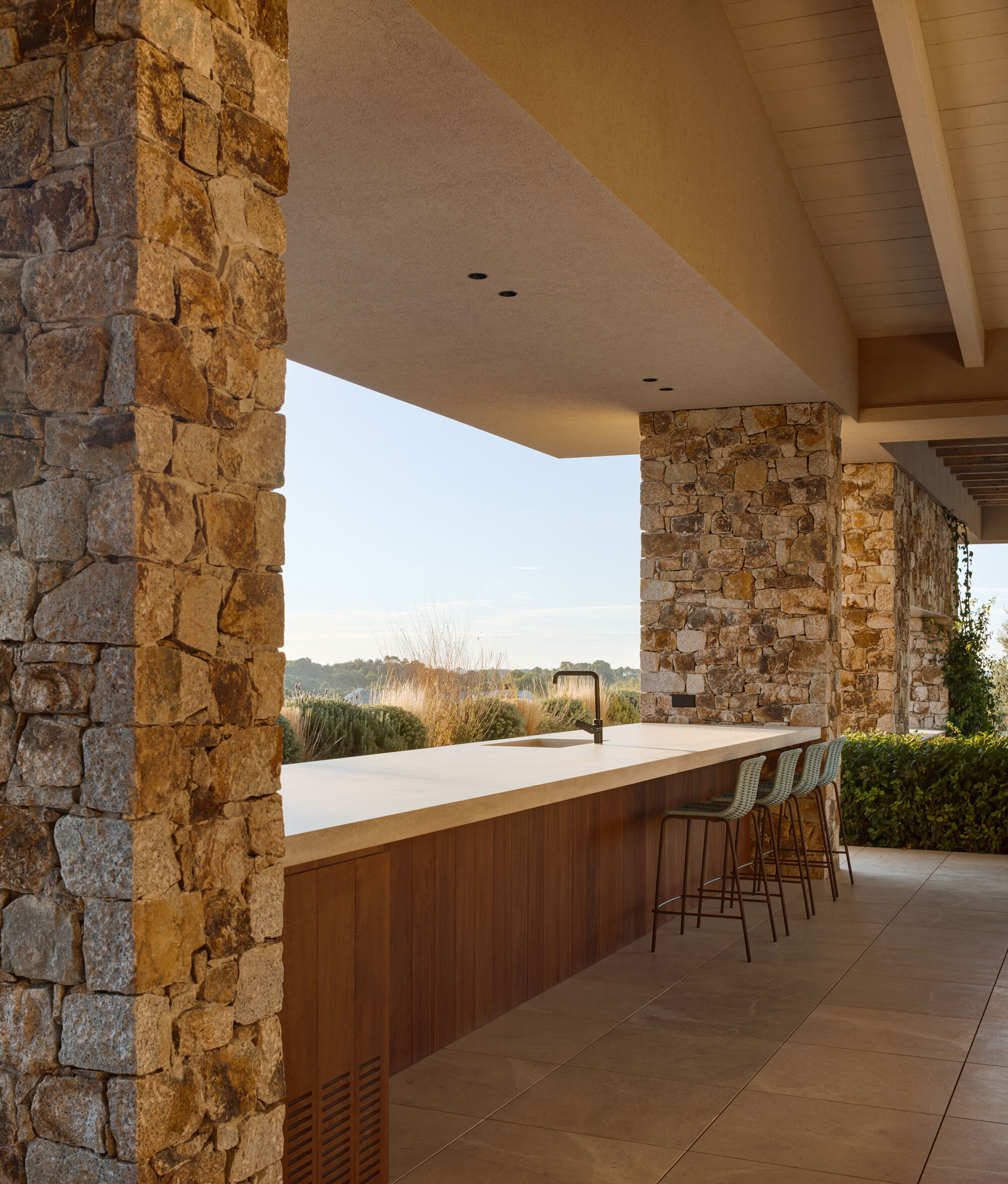 Outdoor kitchen counter with a black tap and high stools between stone columns, facing grasses in soft evening light