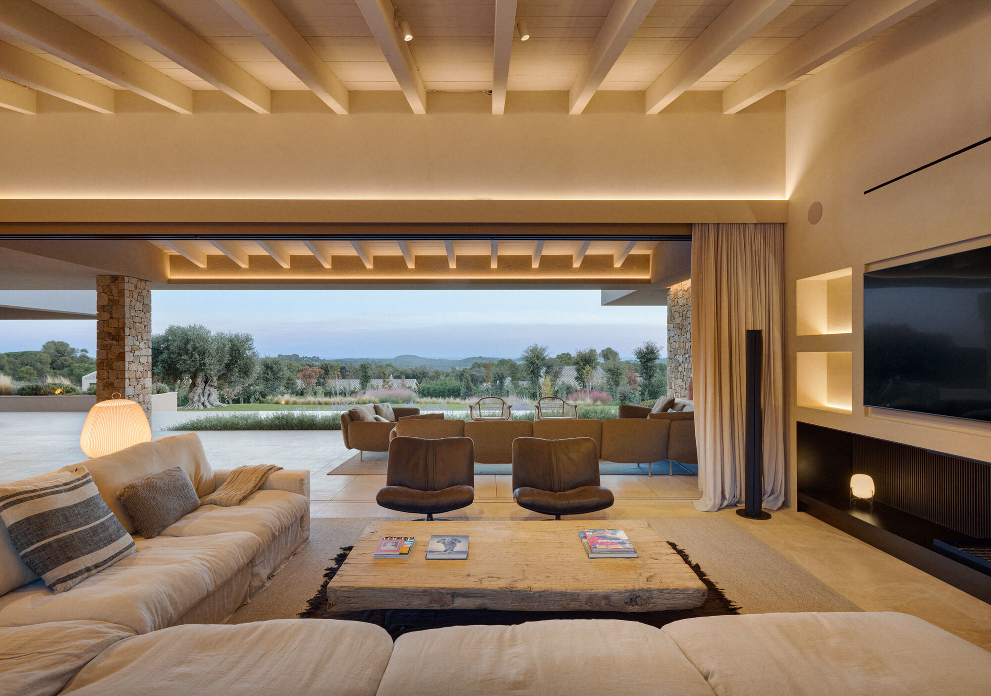 Living room with a rough timber coffee table and leather armchairs, opening to an olive tree and pool terrace at dusk